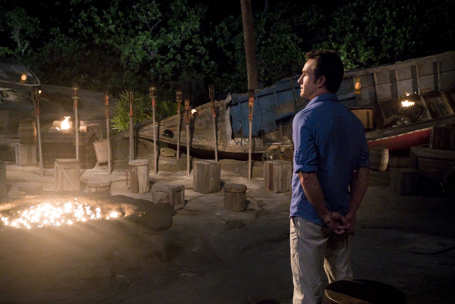 'Survivor' host Jeff Probst wears a blue long-sleeved shirt, rolled up to the elbows, and tan pants. He looks out over Tribal Council.