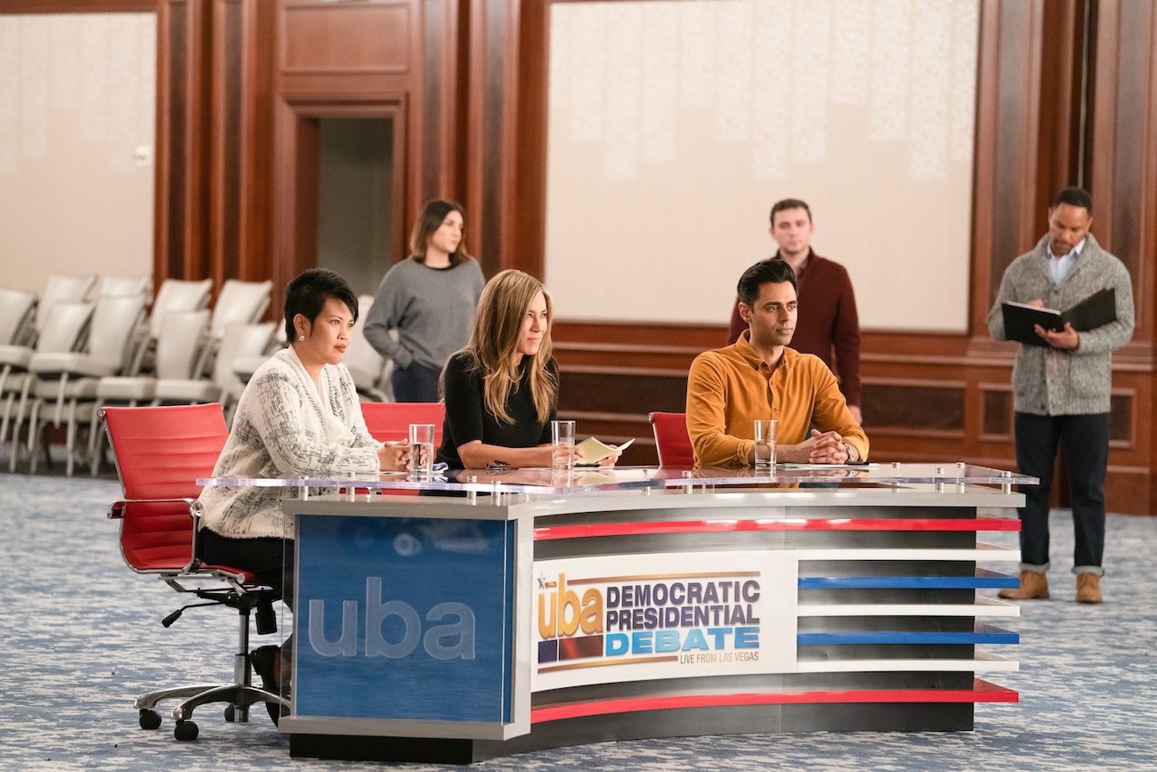 Jennifer Aniston sits at a desk next to Hasan Minhaj and an unnamed woman in a scene from 'The Morning Show' Season 2 Episode 5