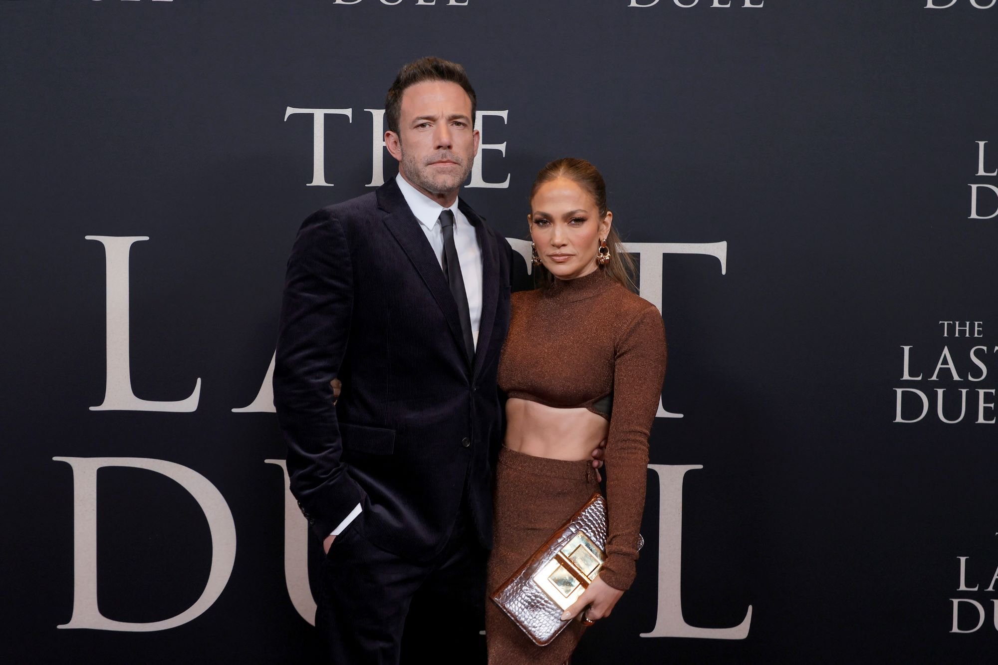 Jennifer Lopez dressed in a brown dress with a gold belt and Ben Affleck dressed in a black suit with a white shirt and a black tie in front of a black background with ivory writing.
