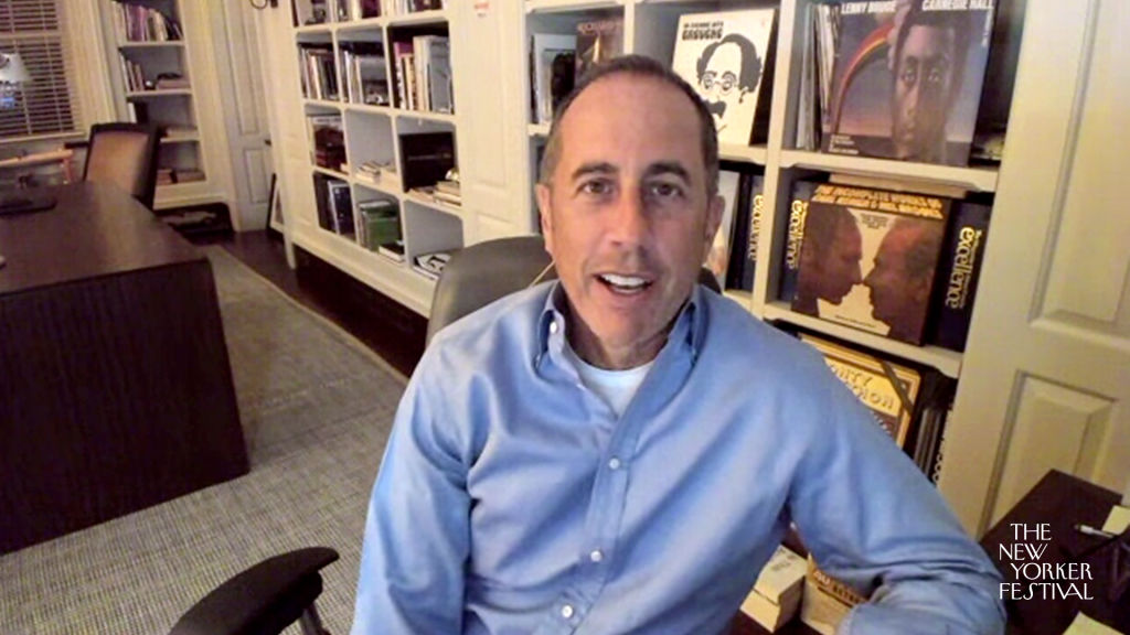Jerry Seinfeld talks into the camera. He's wearing a long-sleeved blue shirt and a bookshelf is behind him.