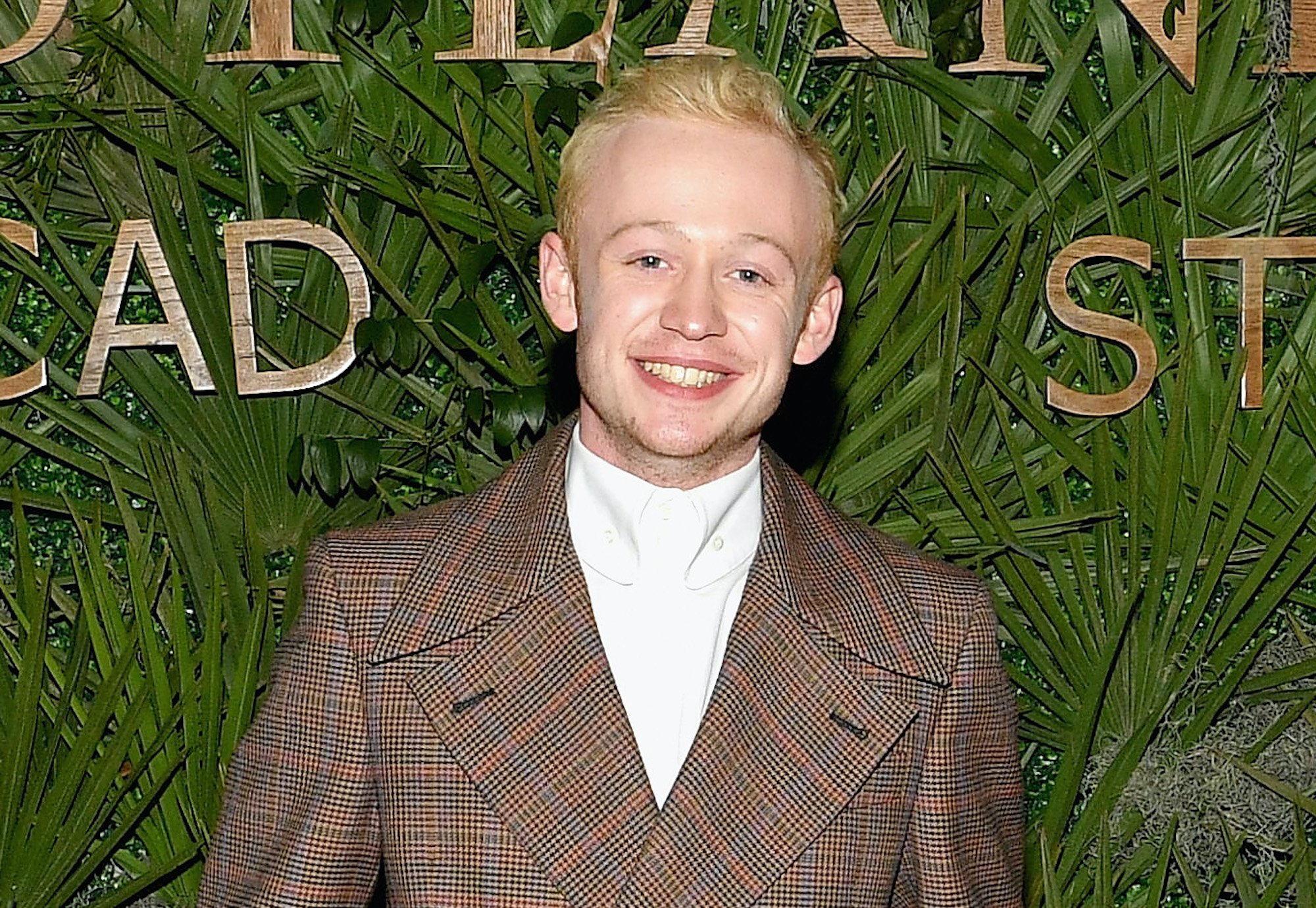 'Outlander' actor John Bell attends the 21st SCAD Savannah Film Festival on October 28, 2018 in Savannah, Georgia. He stands in a brown checkered suit jacket and navy blue pants and smiles in front of a wall covered in green foliage.