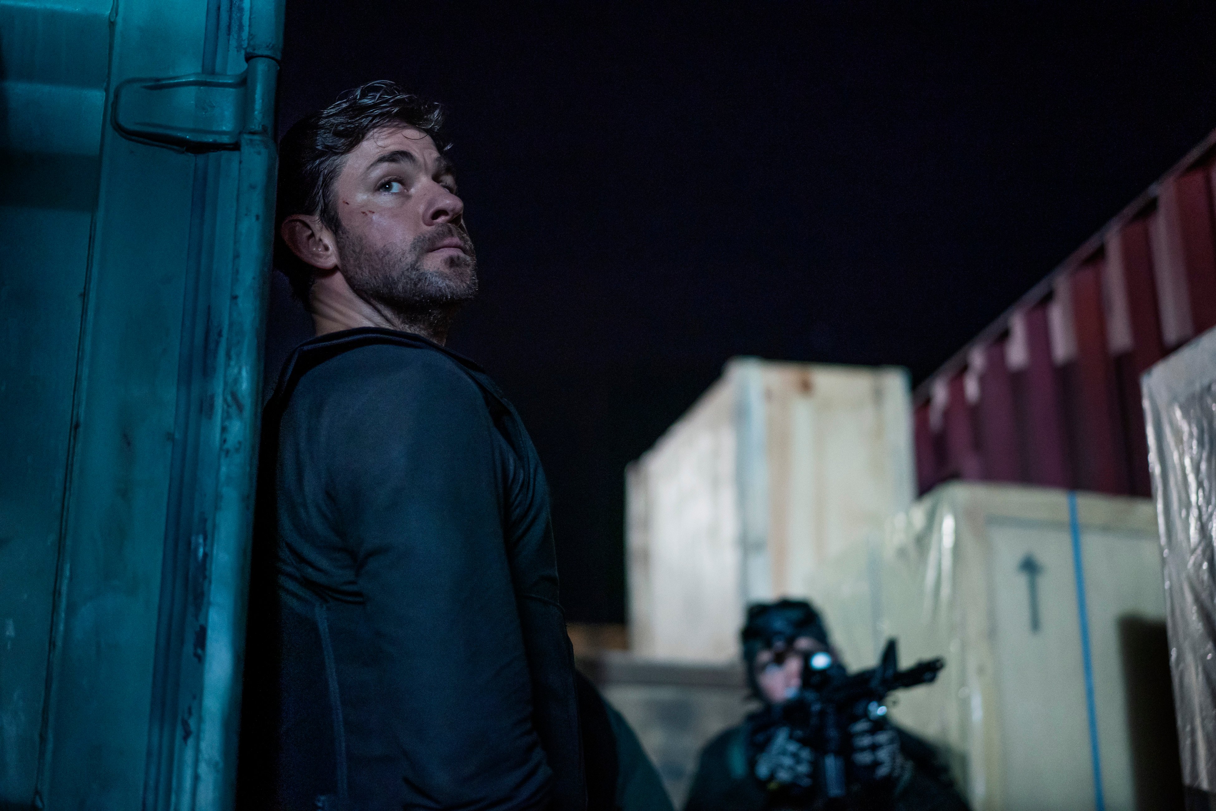 Jack Ryan (John Krasinski) leaning against a wall with a man with a gun in the background in 'Jack Ryan' Season 2