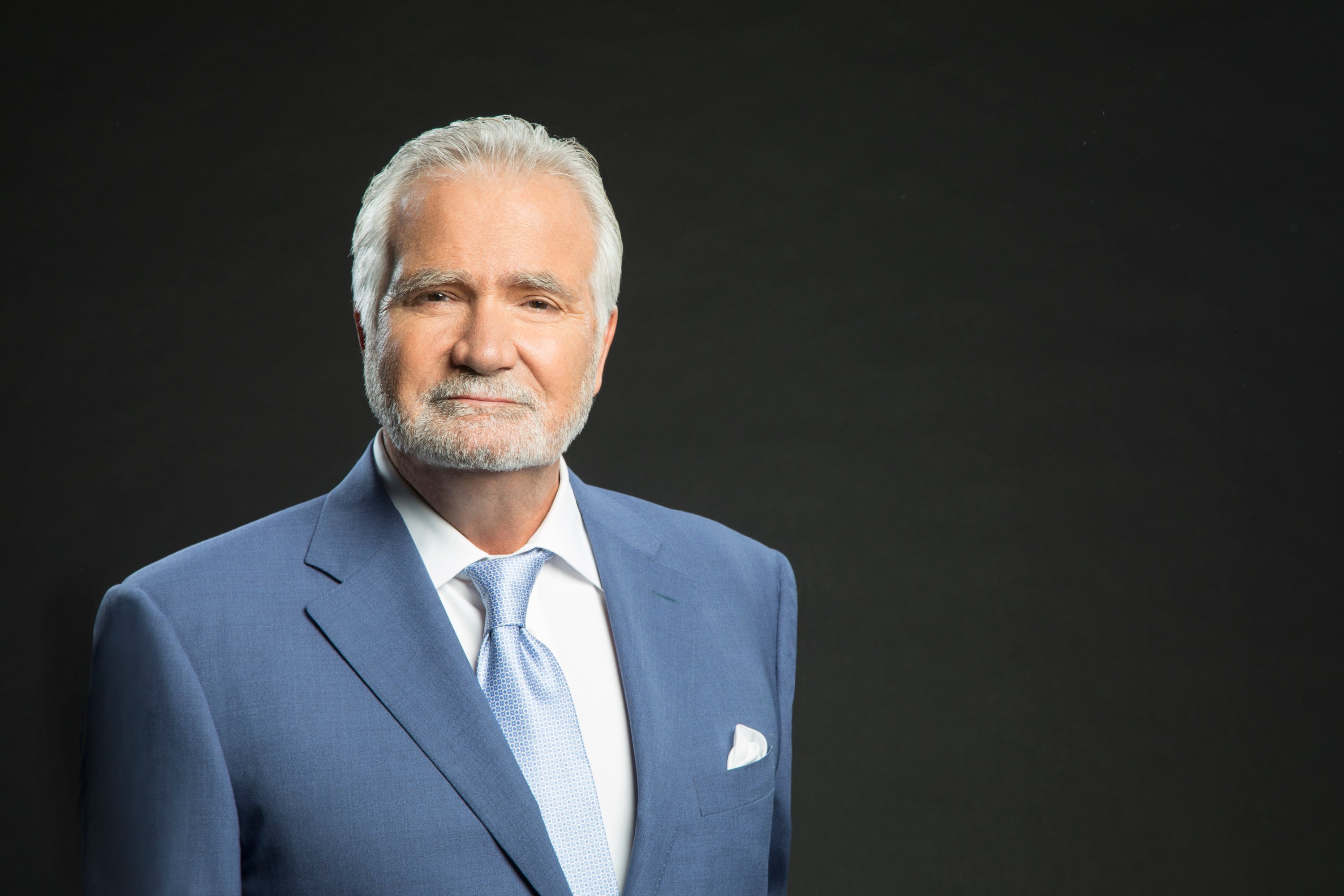'The Bold and the Beautiful' actor John McCook in a white shirt and blue suit and tie standing in front of a black backdrop.