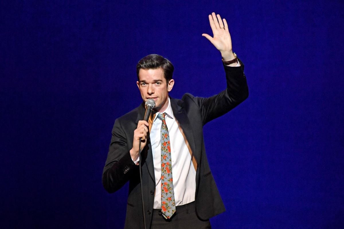 John Mulaney waving in a black suit and floral tie while speaking into a microphone.
