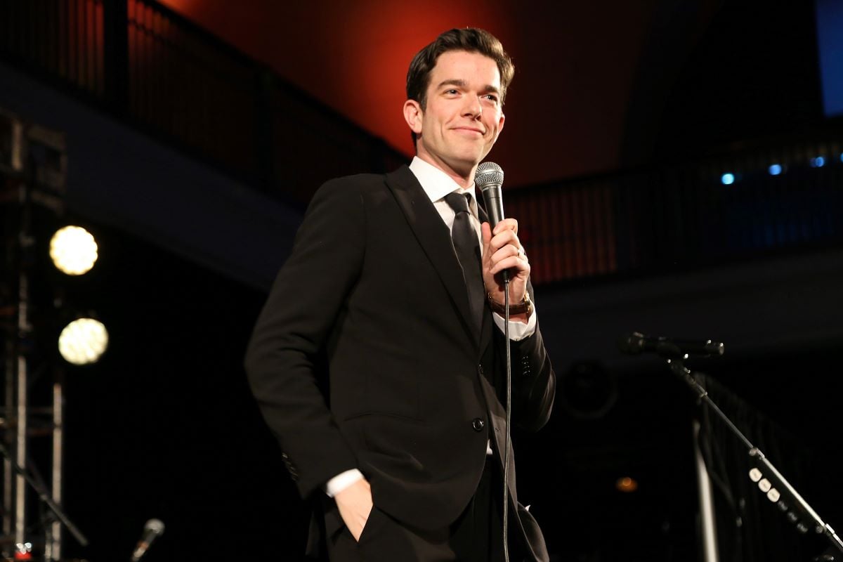John Mulaney on stage in a black suit and tie, holding a microphone