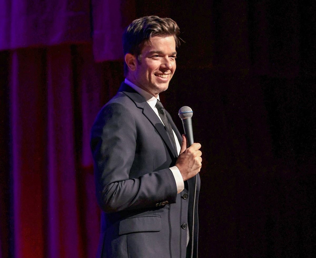 John Mulaney holds a mic, on stage and smiling at audience