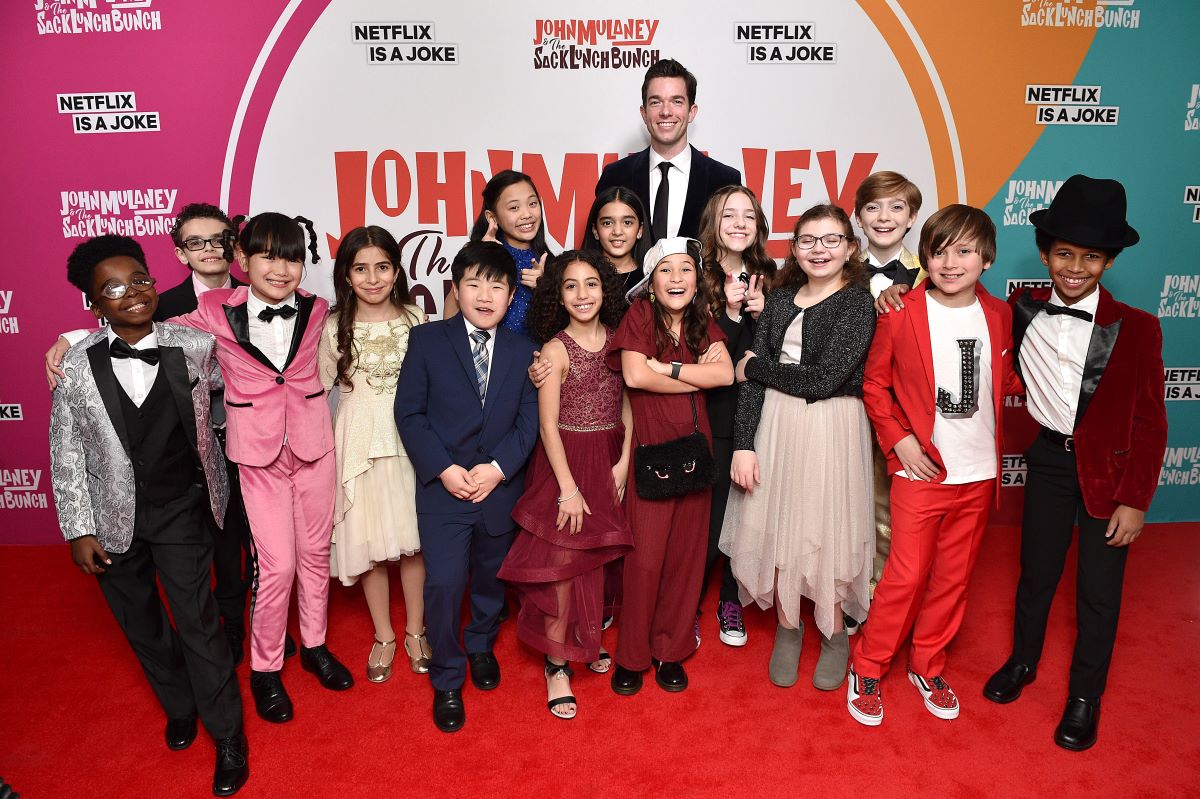 John Mulaney in a black suit and tie surrounded by cast of 'The Sack Lunch Bunch'