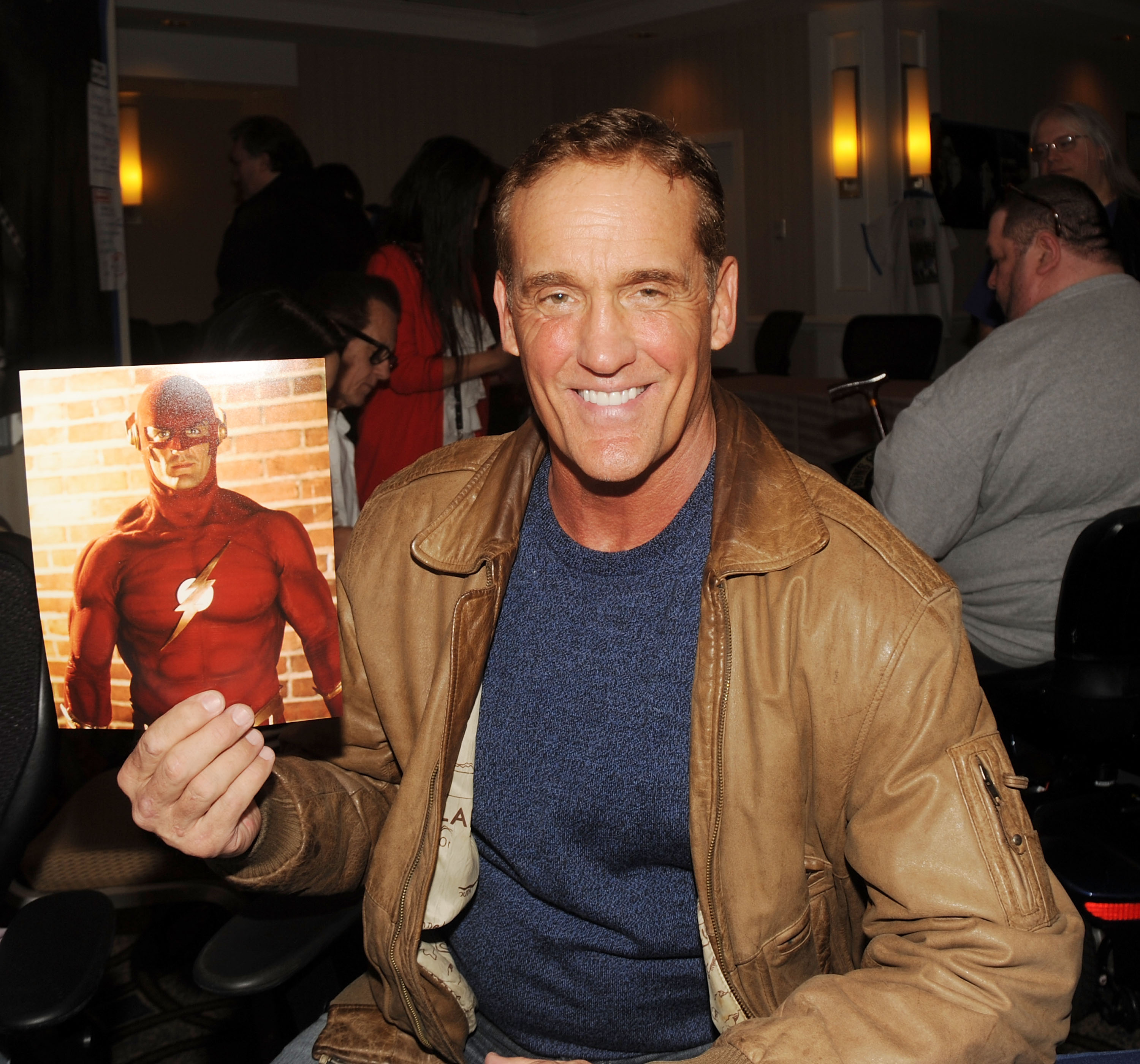 'Stargirl' actor John Wesley Shipp, who plays Jay Garrick, wears a brown jacket over a blue shirt and holds up a picture of him in character as The Flash.
