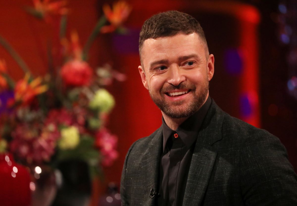 Justin Timberlake, known to an average car, dressed in a black suit with a black shirt in front of a red background with a floral arrangement off to the side.