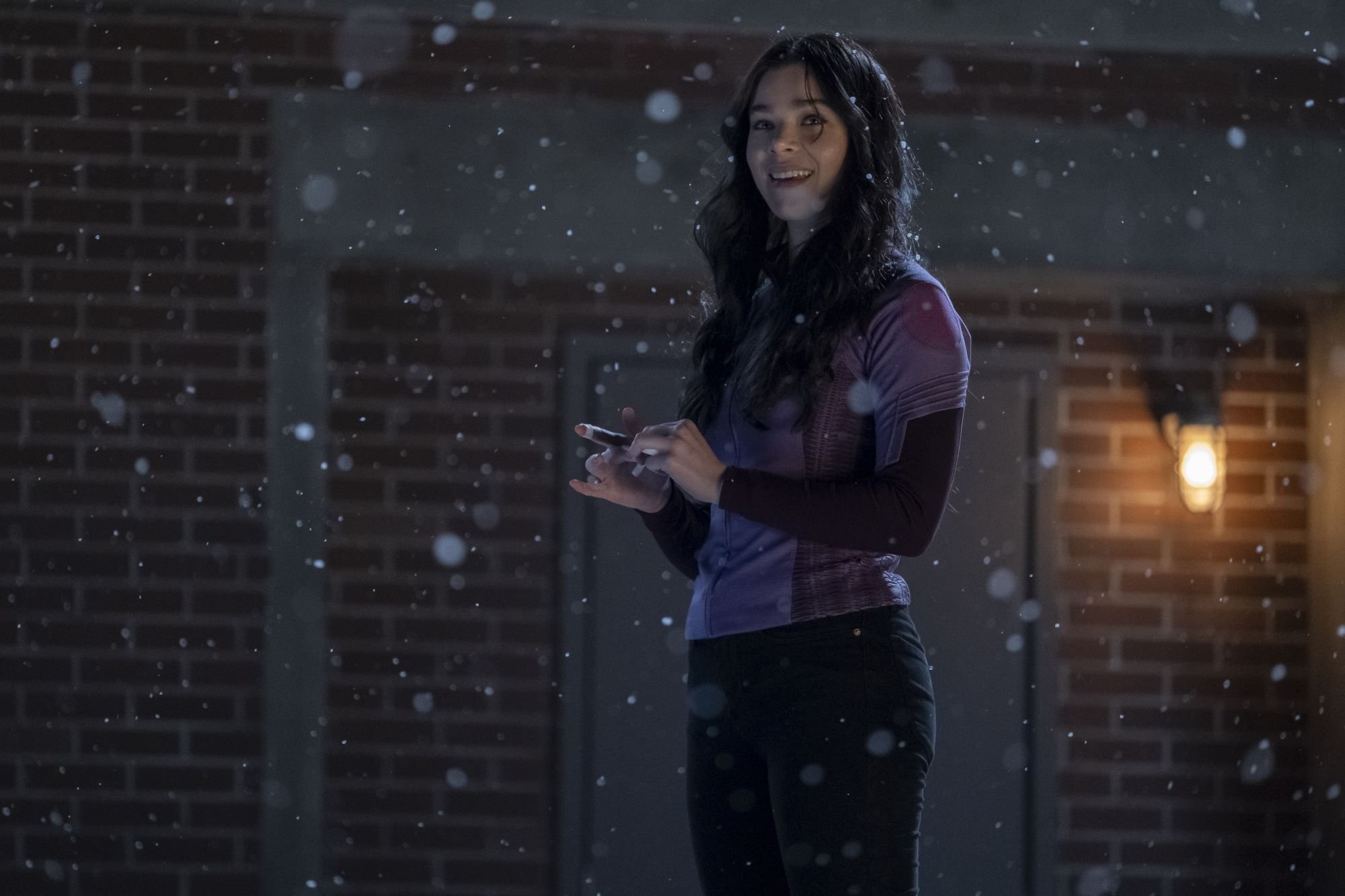 'Hawkeye' star Hailee Steinfeld, in character as Kate Bishop, wears a purple and black long-sleeved shirt and black pants. She smiles as snow comes down around her. Steinfeld can be seen in the new 'Hawkeye' trailer.