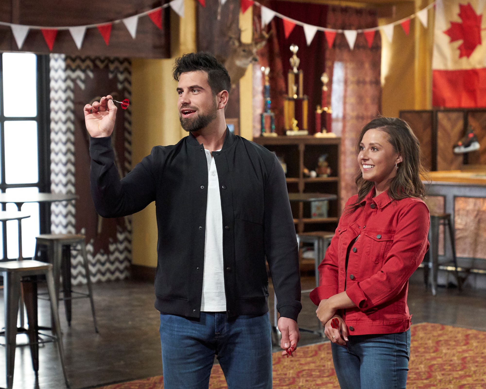 'The Bachelorette' stars Blake Moynes and Katie Thurston play darts. Blake wears a black jacket over a white shirt and jeans. Katie wears a red jacket and jeans.