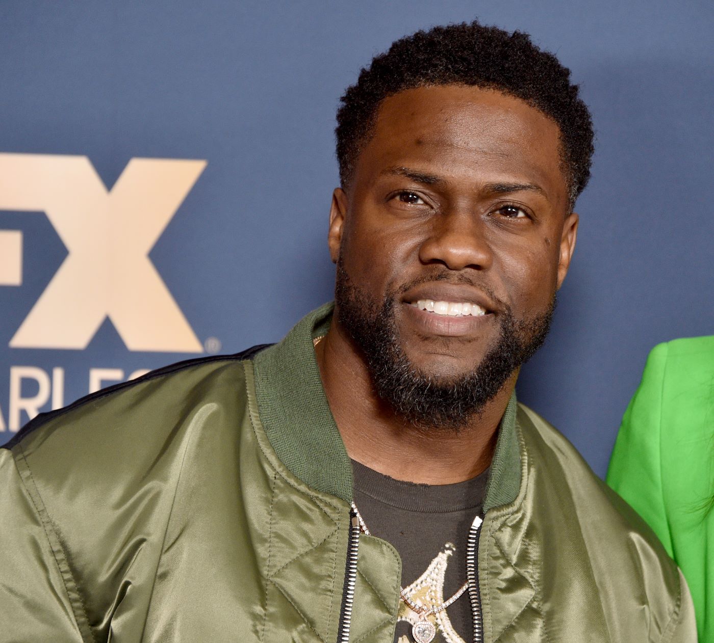 Kevin Hart dressed in a olive green shiny jacket with a darker green underneath in front of a blue background.