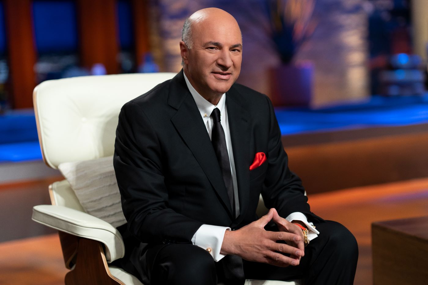 Kevin O'Leary sitting a chair on the set of 'Shark Tank' in a traditional black suit with a white shirt and black tie.