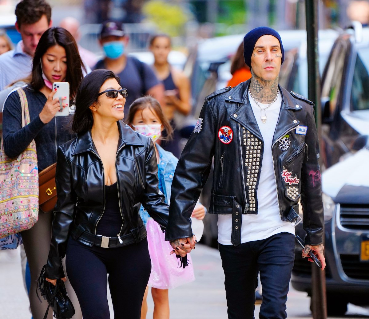 Kourtney Kardashian and Travis Barker smile while walking hand-in-hand down a busy street.