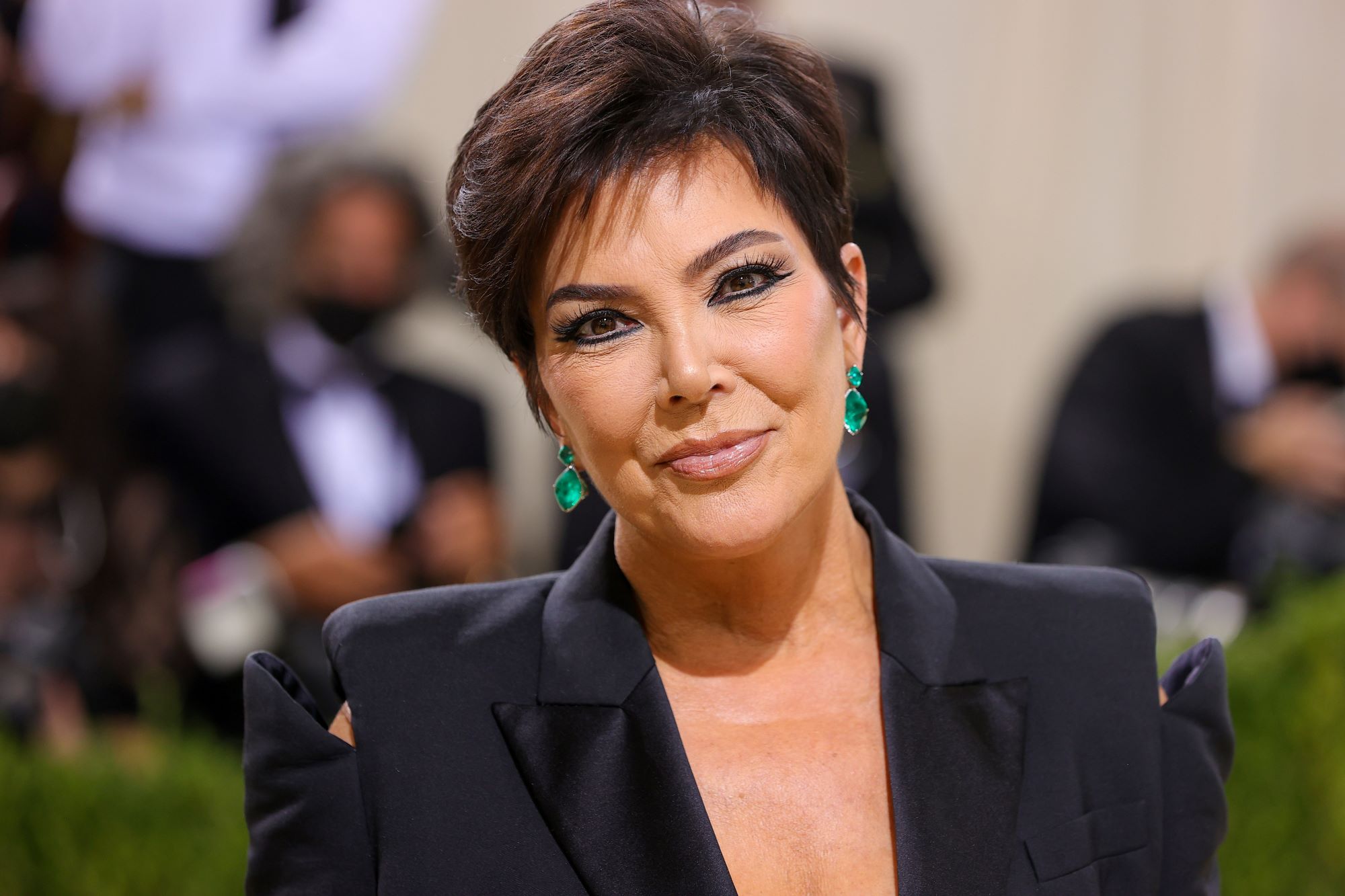 Kris Kardashian dressed in a black suit jacket with a blurred background of various people.