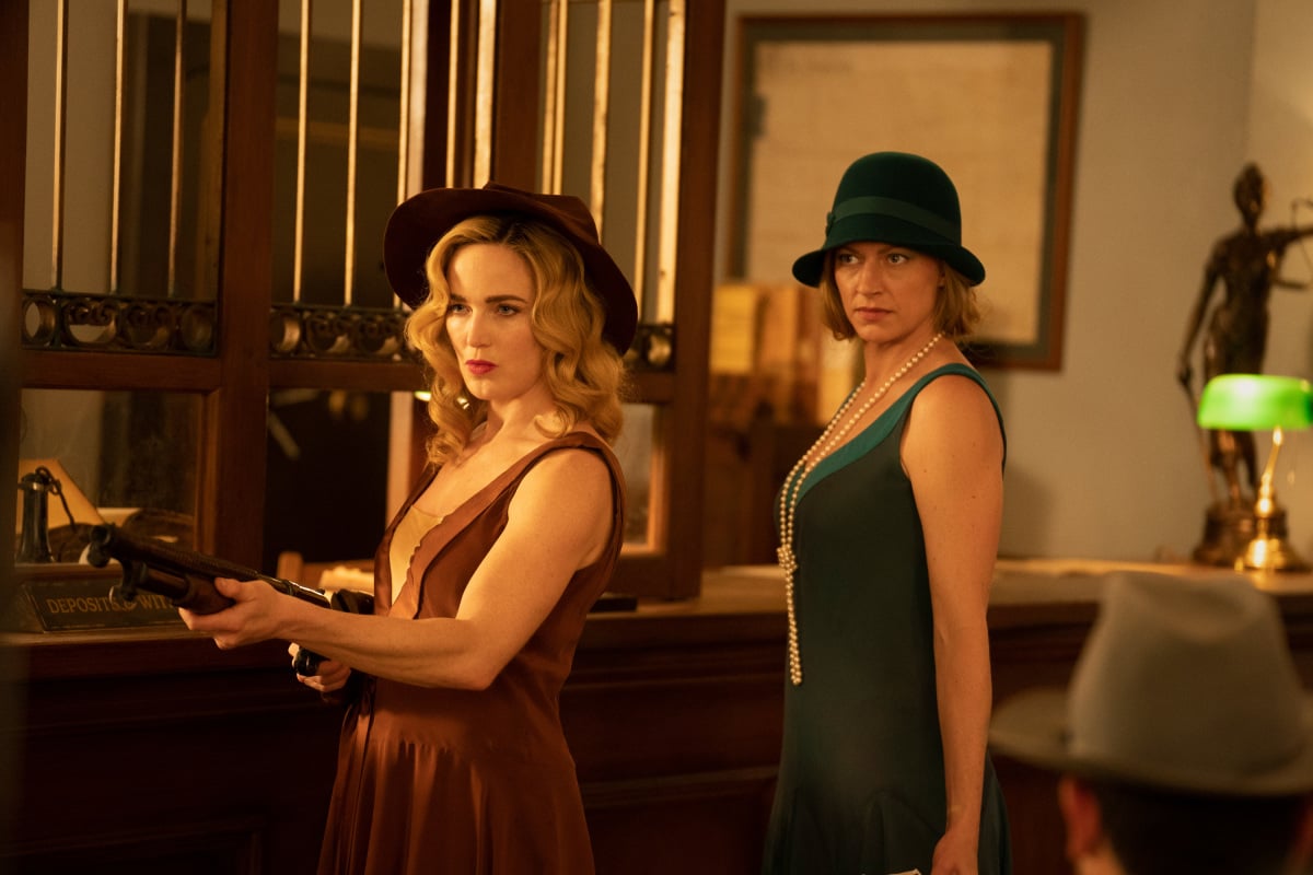 'DC's Legends of Tomorrow' Season 7 Episode 1 actors Caity Lotz and Jes Macallan dress in '20s outfits. Lotz wears a brown dress and a brown hat while wielding a gun. Macallan wears a green dress, green hat, and a set of long pearls.