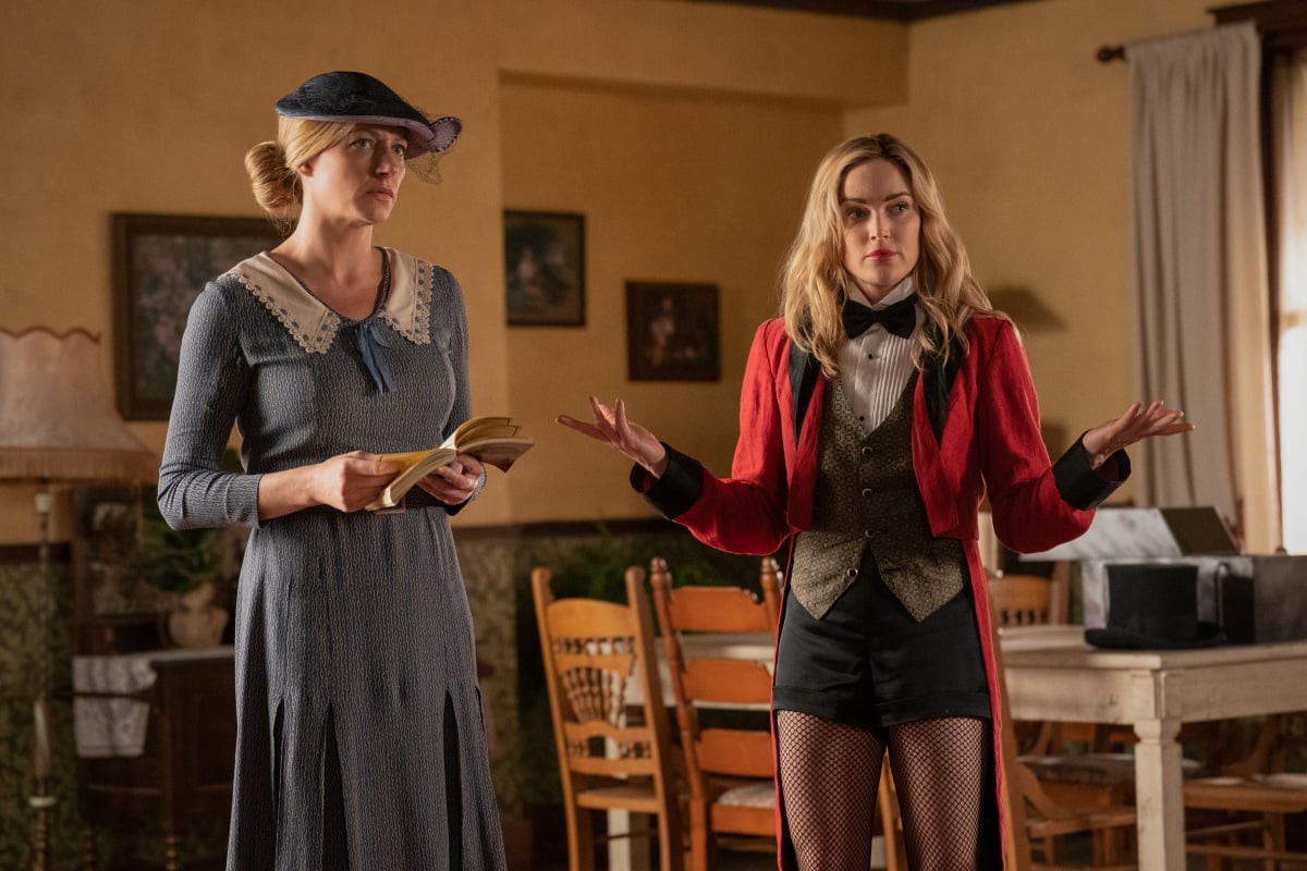 'DC's Legends of Tomorrow' Season 7 stars Jes Macallan and Caity Lotz, in character as Ava Sharpe and Sara Lance. Ava wears a blue dress and blue hat. Sara wears a red magician's jacket over a white shirt, green vest, and bow tie.