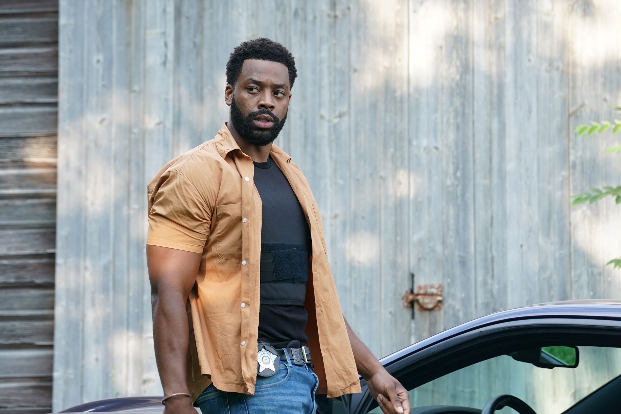 LaRoyce Hawkins playing Kevin Atwater in 'Chicago P.D.' wearing a brown open button-up T-shirt with a black shirt and blue jeans leaning up against a car in front of a grey wall.