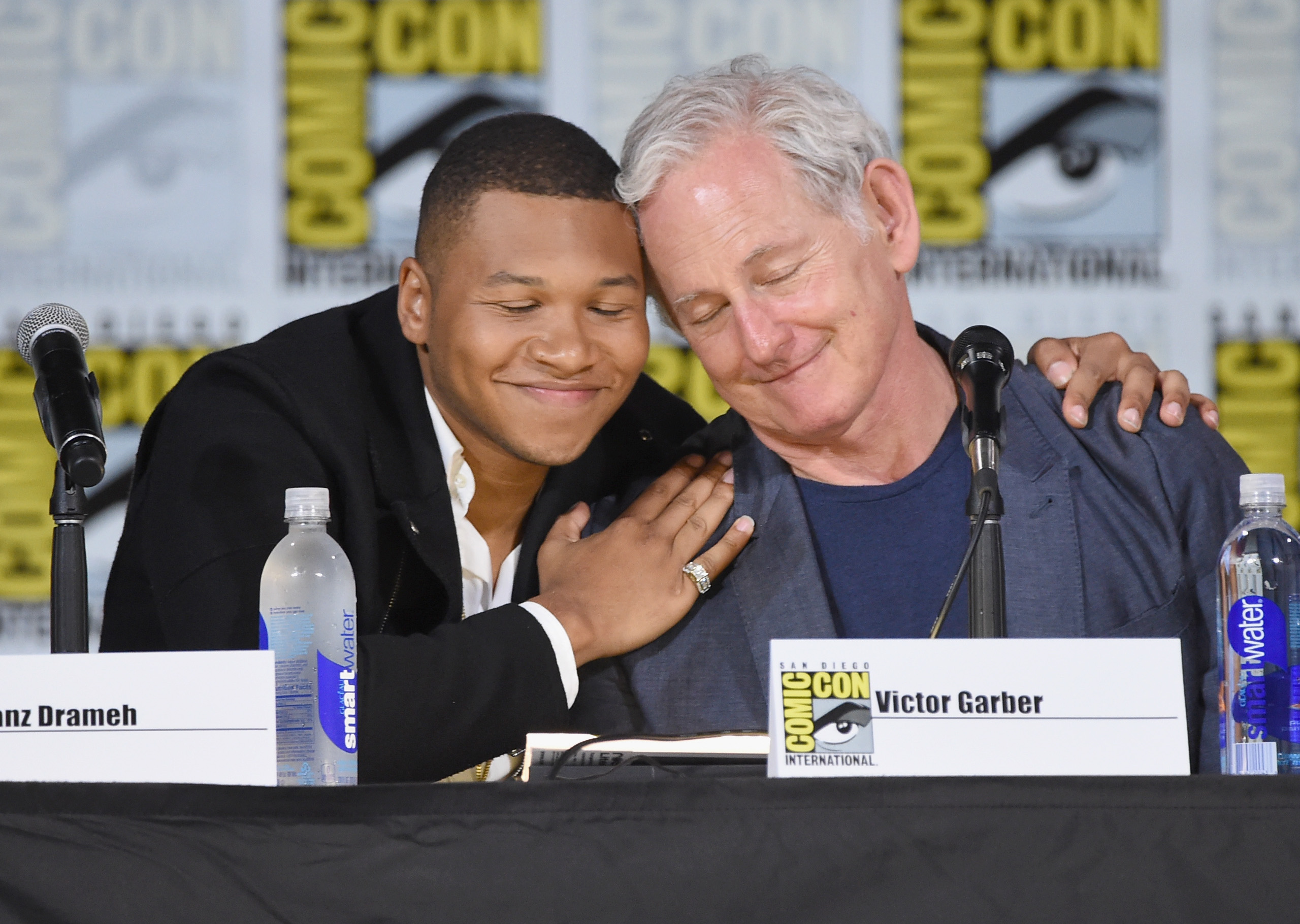 'DC's Legends of Tomorrow' actors Franz Drameh and Victor Garber embrace onstage during a panel at San Diego Comic-Con. Drameh wears a black suit over a white shirt. Garber wears a blue suit over a blue shirt.