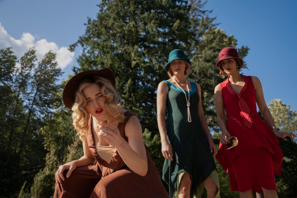 'Legends of Tomorrow' stars Caity Lotz, Jes Macallan, and Tala Ashe, in character as Sara, Ava, and Zari, look at something on the ground. Lotz wears a brown dress and brown hat. Macallan wears a green dress and green hat. Ashe wears a red dress and red hat. 'Legends of Tomorrow' is streaming on Netflix.