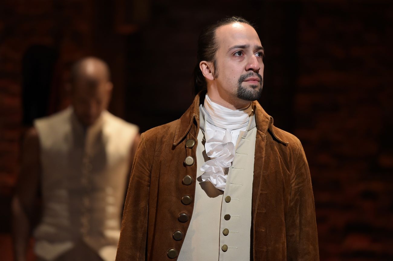 Lin-Manuel Miranda wears a tan best, ruffled collar, and brown coat in a performance of Hamilton.