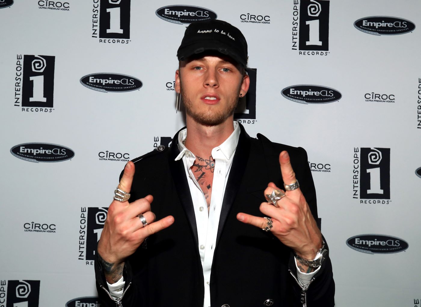 Machine Gun Kelly in a black suit with a white shirt in front of a white background with writing on it.