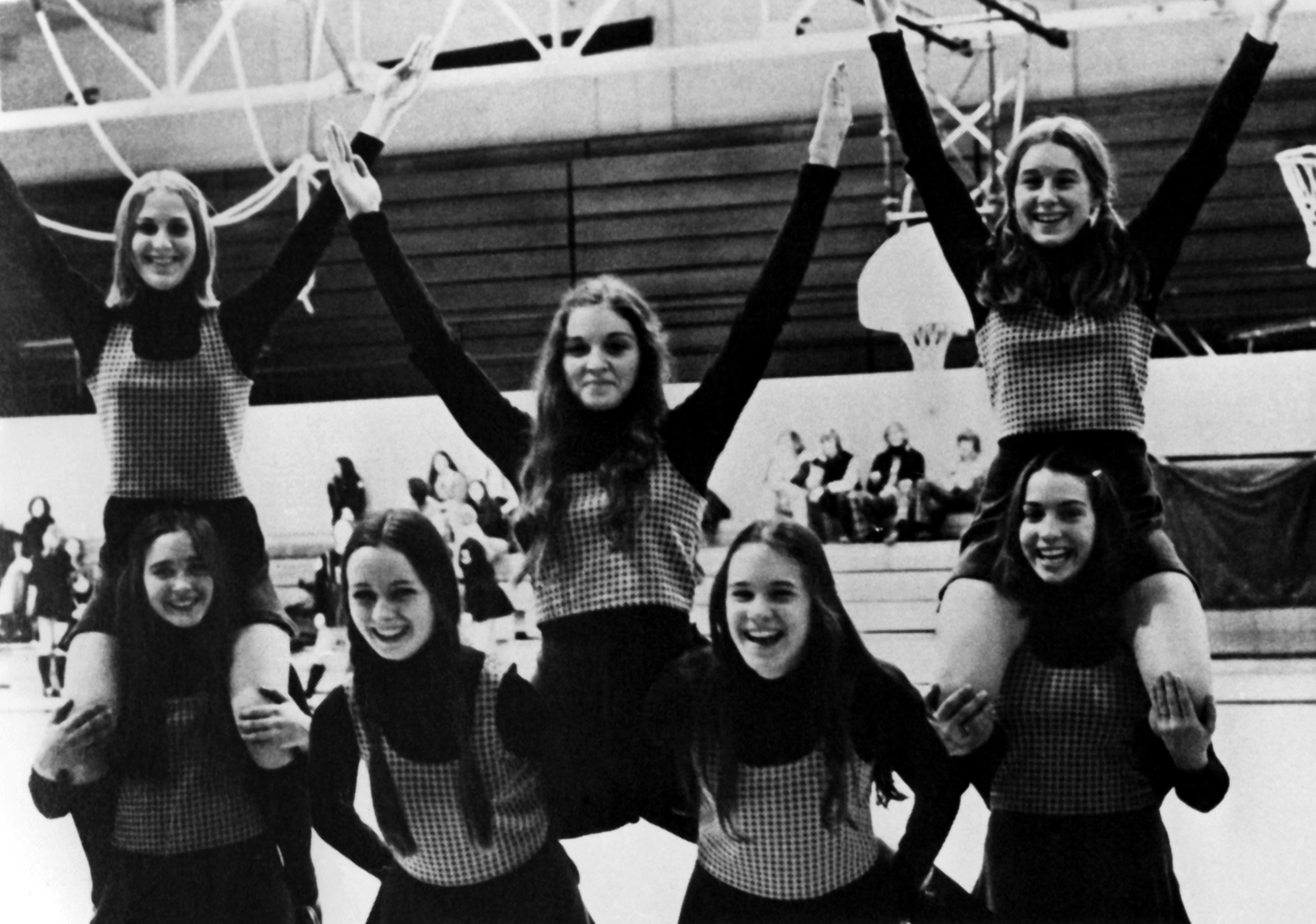 A black and white photo of Madonna and other high school cheerleaders. She holds her hands up in the air.