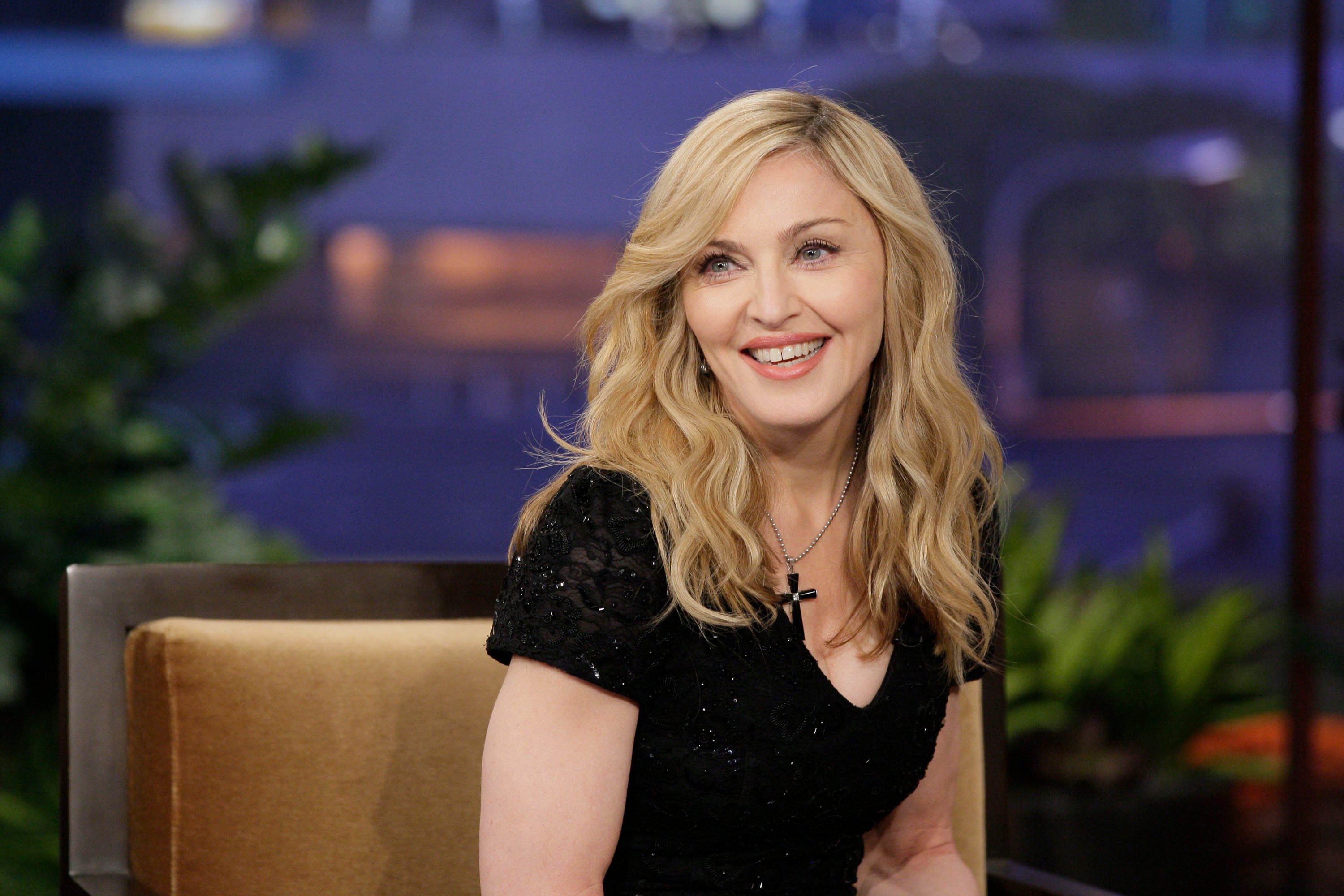 Madonna wears a black shirt and black cross necklace. She sits in a chair.