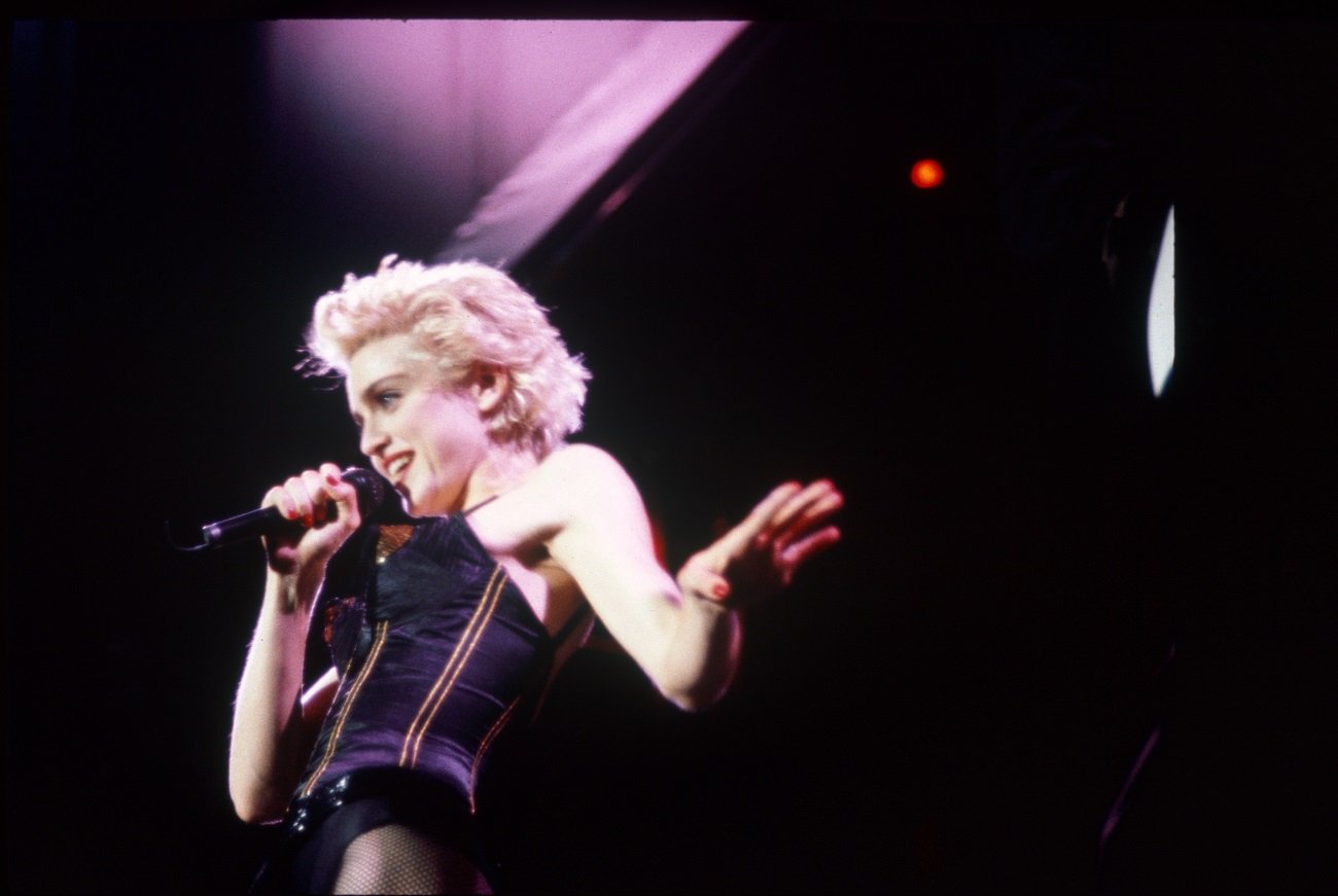 Madonna wears a black top and fishnet tights. She holds a microphone.