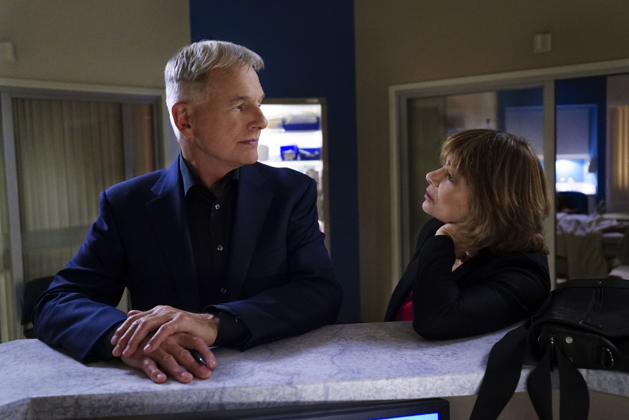 Mark Harmon wears a dark jacket while standing at a desk with Laura San Giacomo