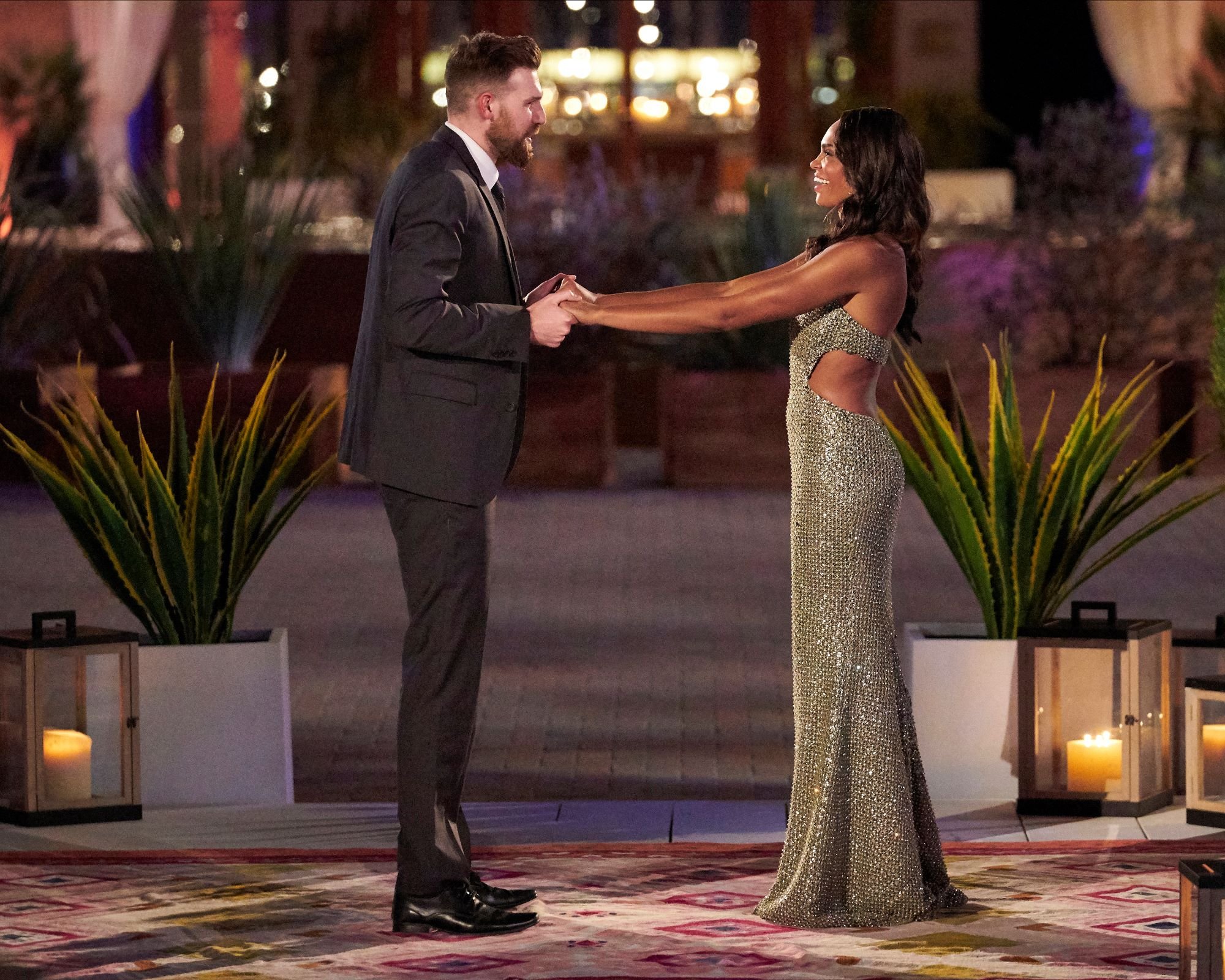 'The Bachelorette' star Michelle Young meets one of her contestants, Chris Gallant, for the first time. Chirs wears a black suit and black dress shoes. Michelle wears a muted green sparkly dress. They hold hands.