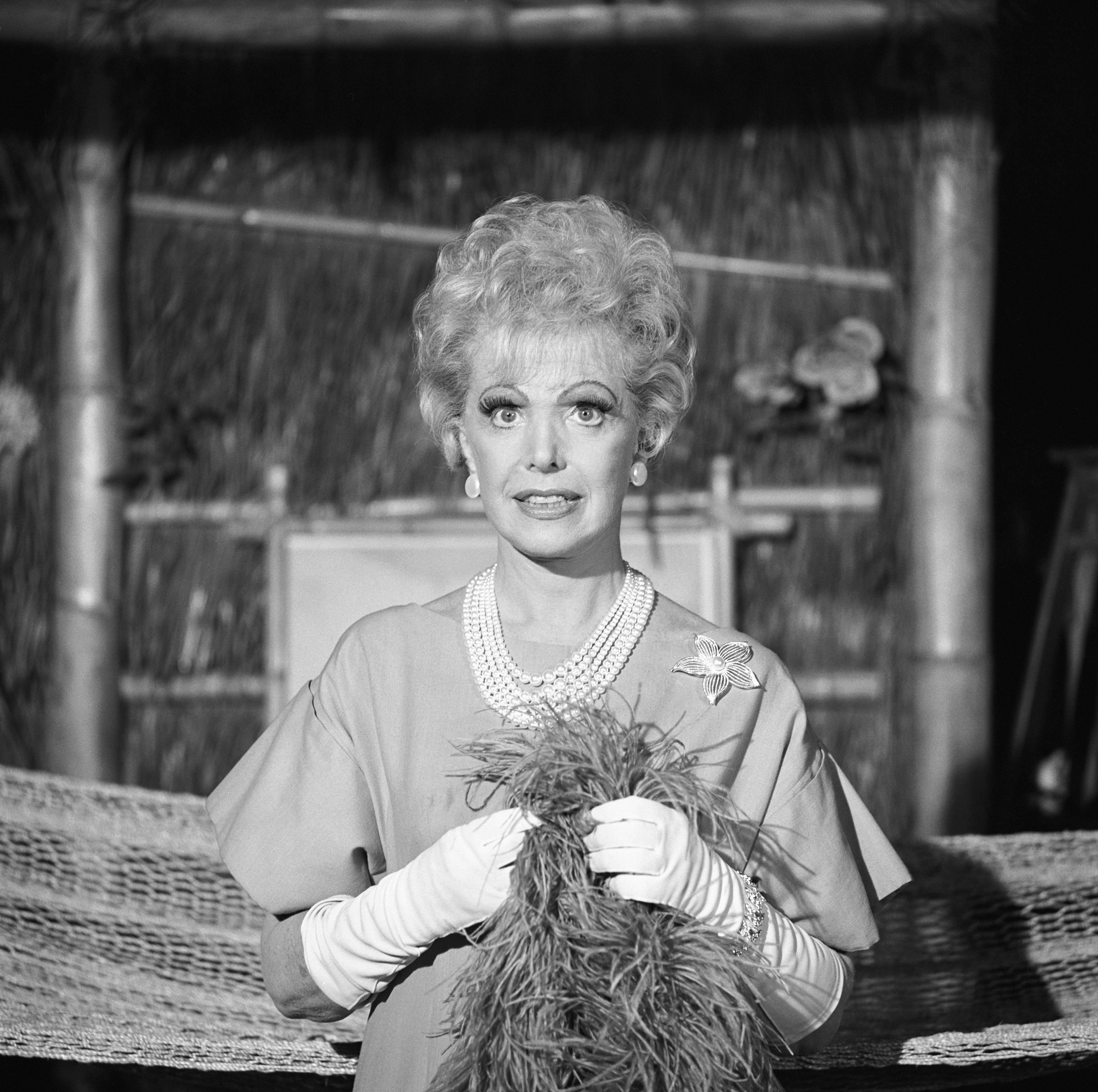 'Gilligan's Island' actor Natalie Schafer wearing a dress, long gloves, pearl necklace, and feather boa standing in front of a bamboo hut.