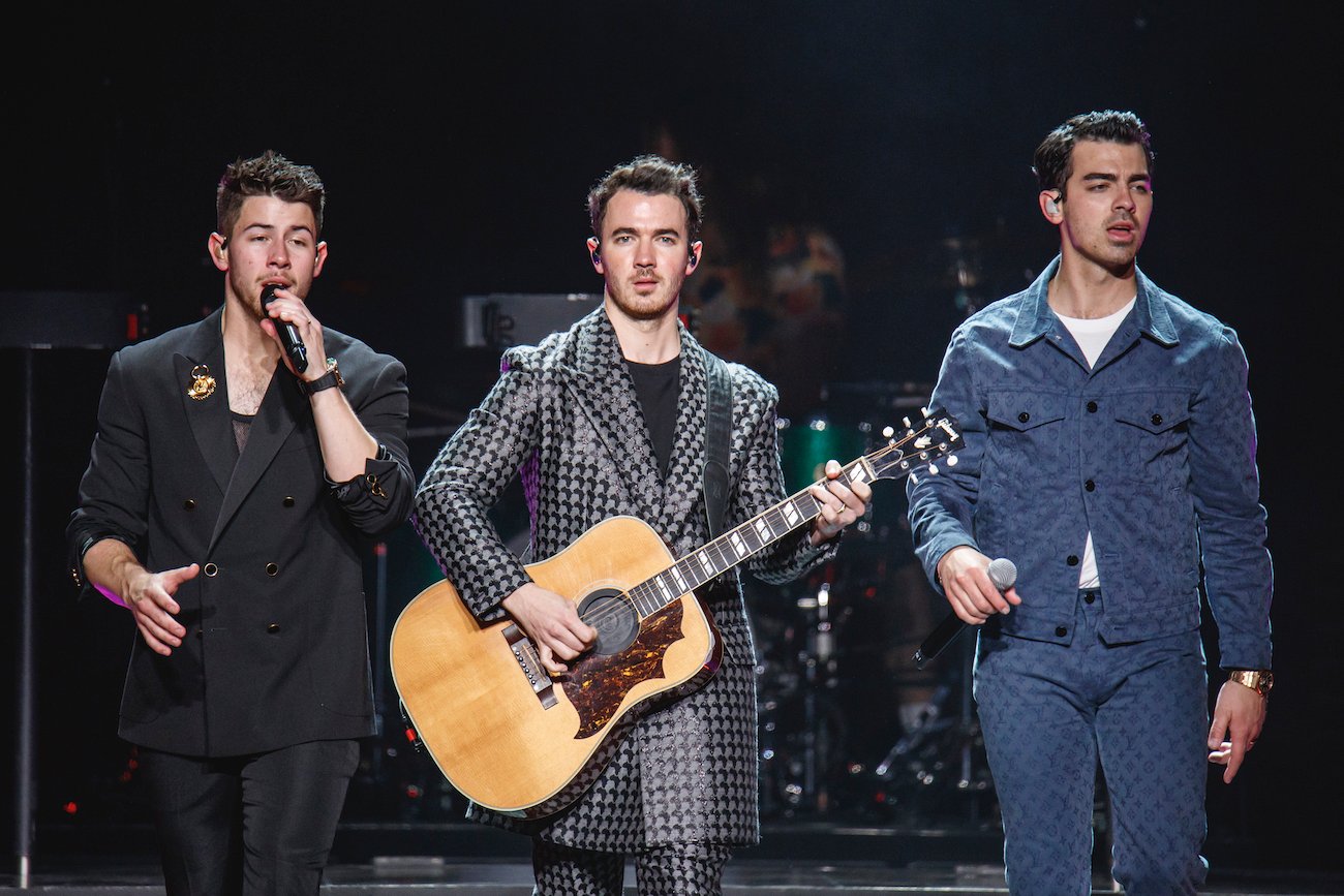 Nick Jonas sings into a mic while Kevin Jonas plays guitar and Joe Jonas looks on