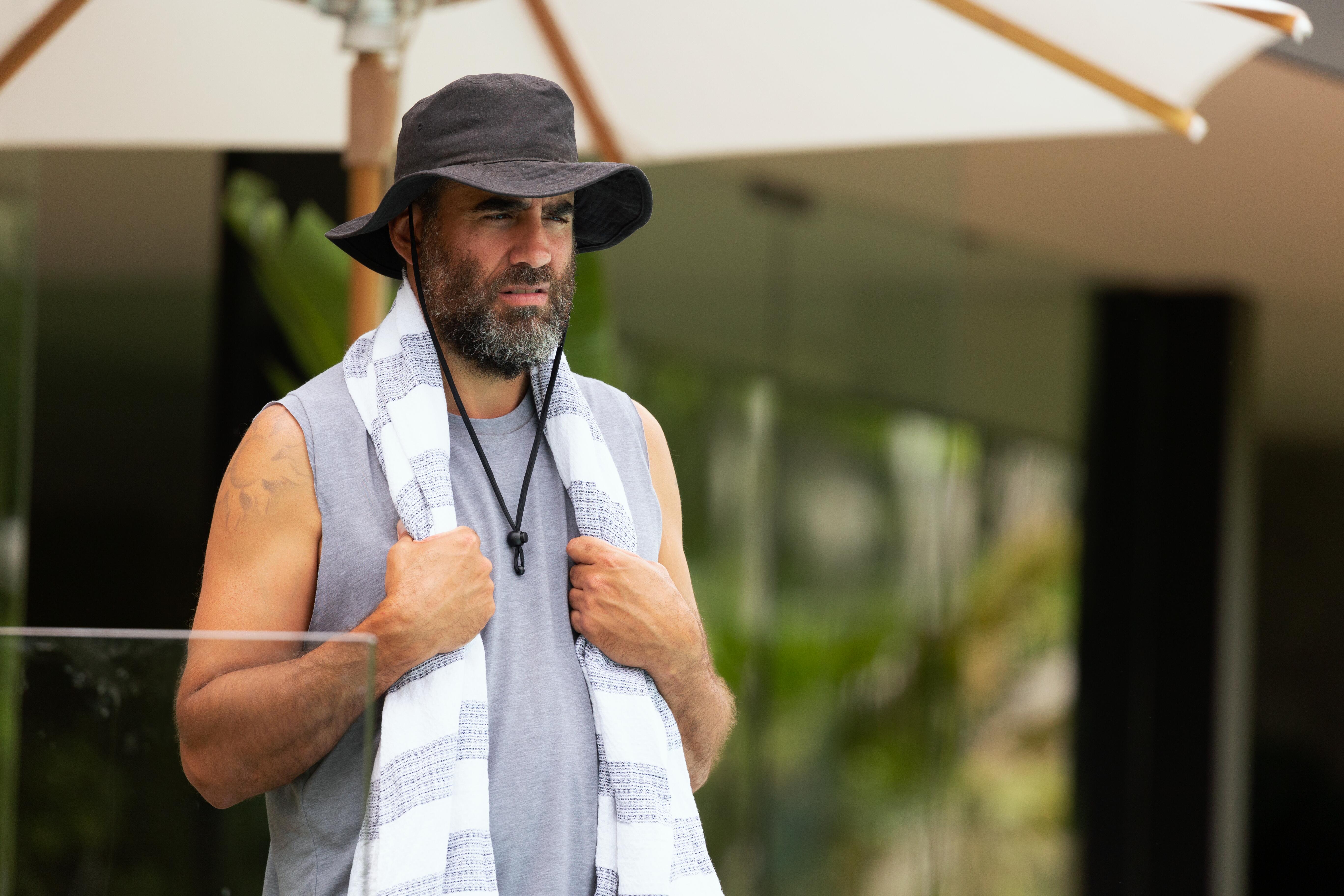 Tony Hogburn stand wearing a grey tank shirt and wide-brimmed hat with a towel around his neck in Nine Perfect Strangers.