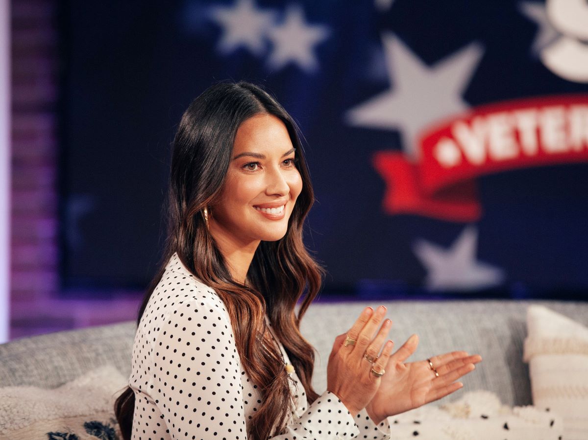 Olivia Munn in a black and white polka dot shirt, sitting and clapping