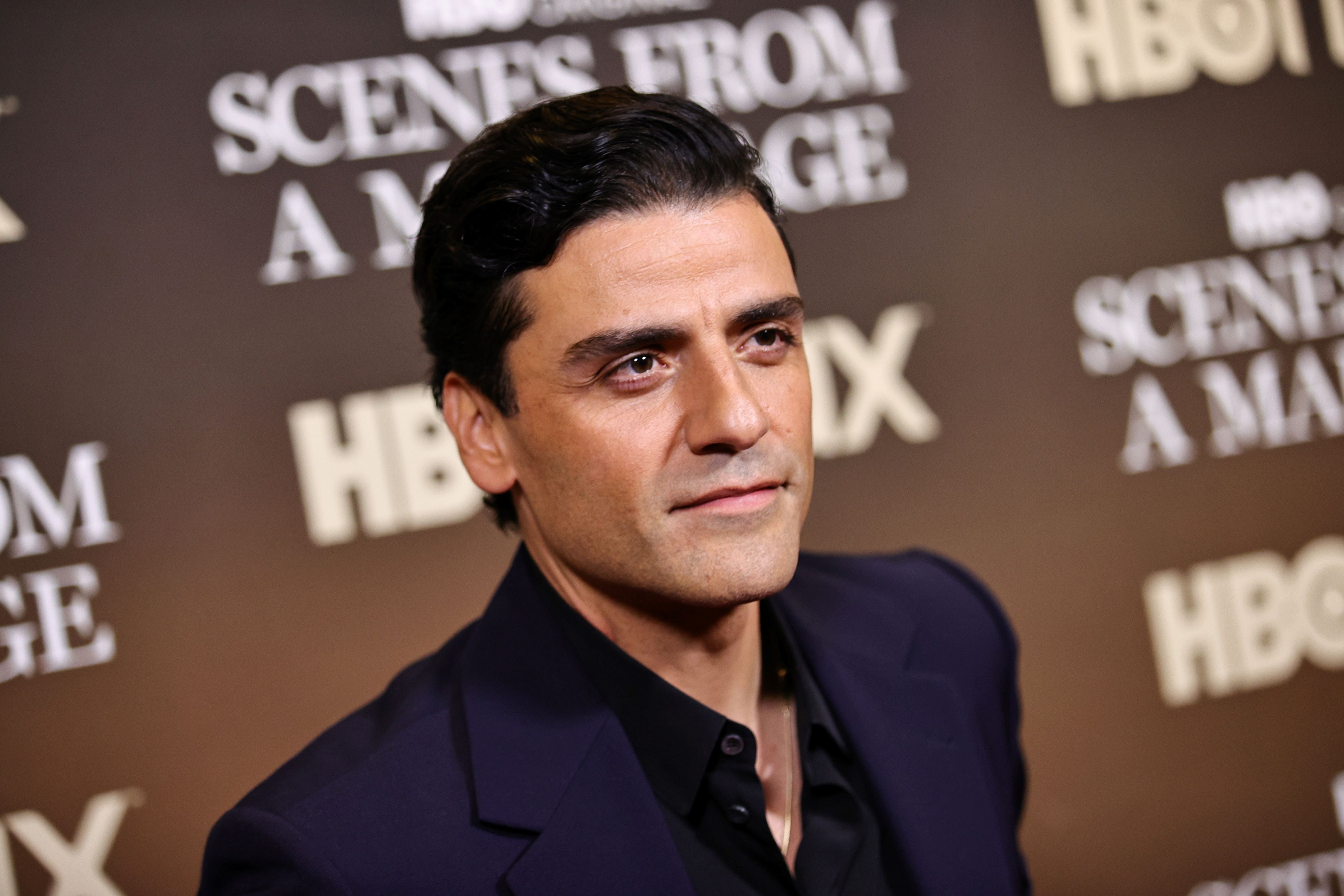 Oscar Isaac standing in front of a brown background with writing on it dressed in black.