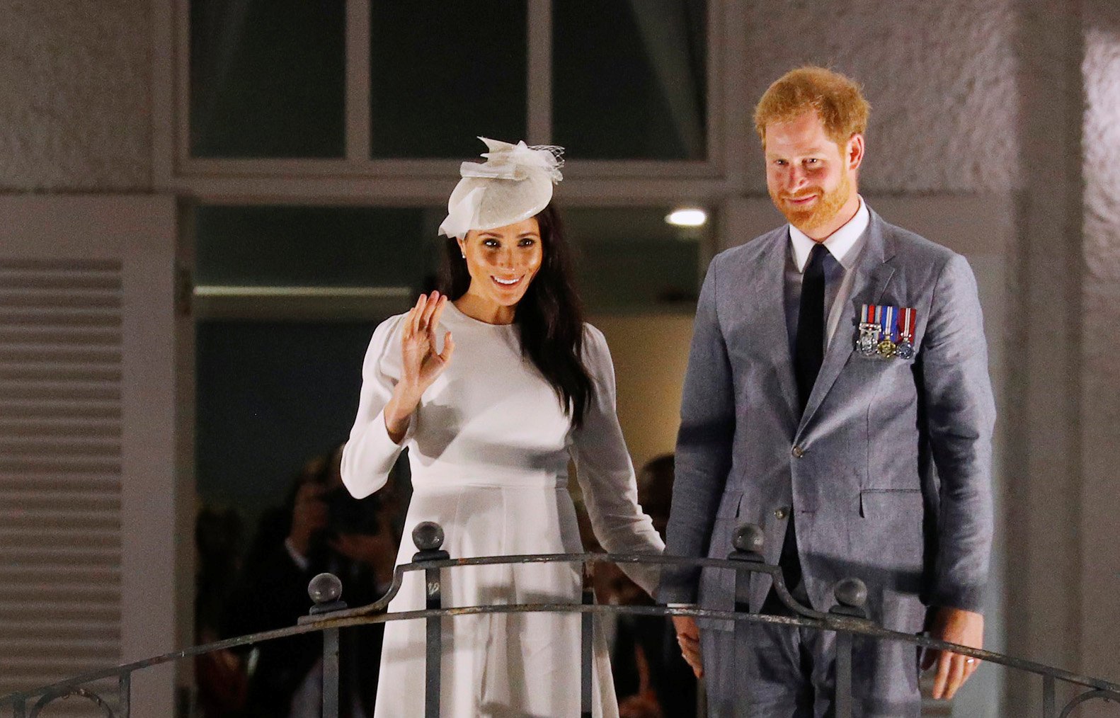 Prince Harry wearing a gray suit and Meghan Markle in a white dress waving from the balcony of the Grand Pacific Hotel in Suva, Fiji