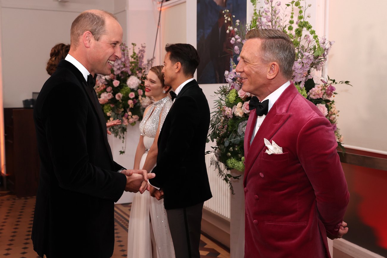 Prince William smiles in a black tuxedo as he looks at a smiling Daniel Craig