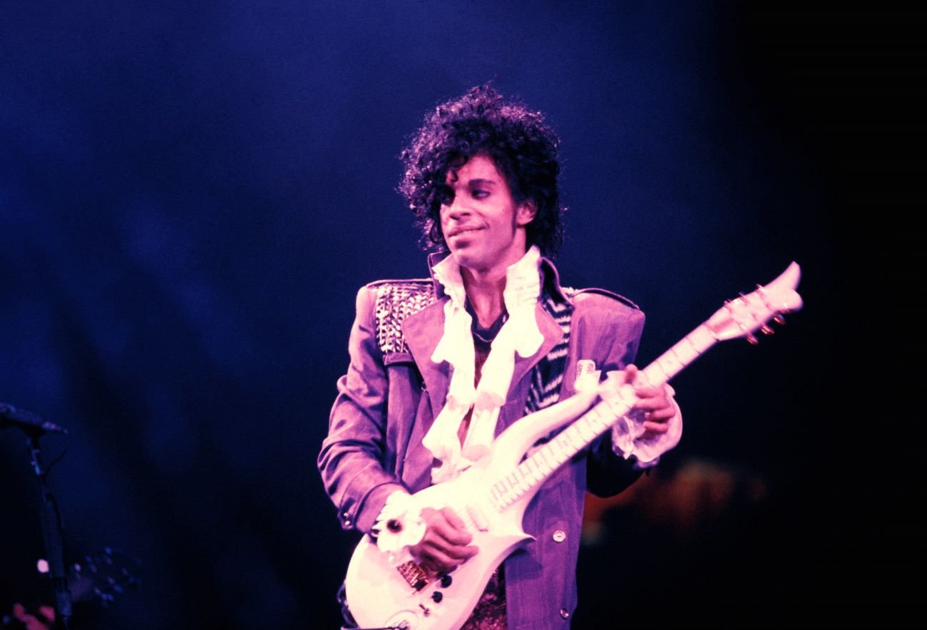 Prince wears a purple jacket and holds a white guitar.