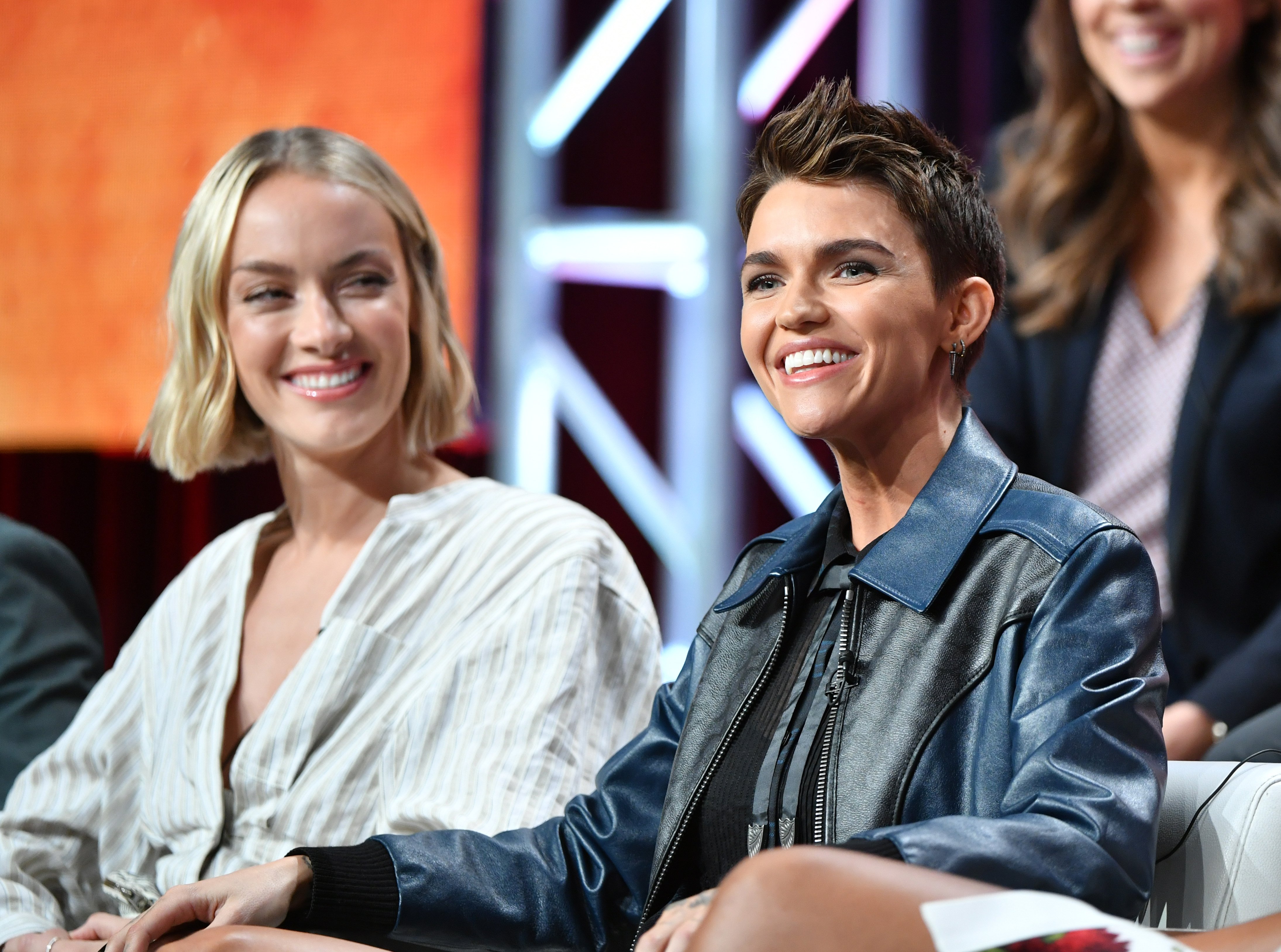 'Batwoman' stars Rachel Skarsten and Ruby Rose sit side-by-side. Skarsten wears a white blouse. Rose wears a blue and black leather jacket.