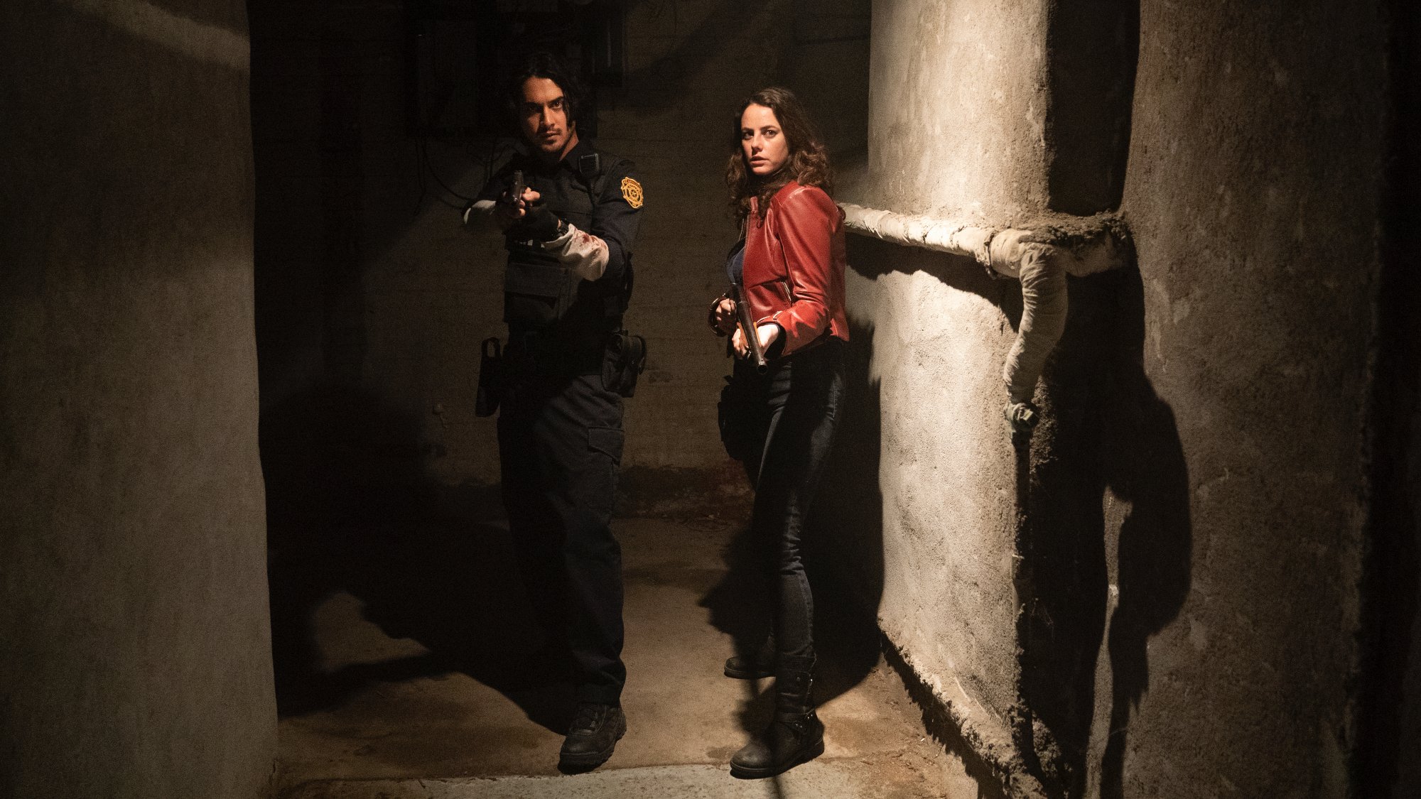 'Resident Evil Welcome to Raccoon City' actors Avan Jogia as Leon Kennedy and Kaya Scodelario as Claire Redfield with guns in a dark hallway