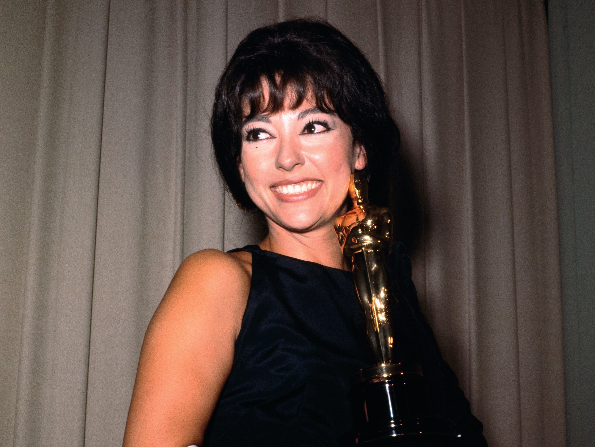 Rita Moreno holding her Academy Award for her performance as Anita in 'West Side Story.' Moreno smiles in a black and gold dress and long black gloves as she holds the golden Oscar statuette in her hands backstage at the 1962 ceremony.