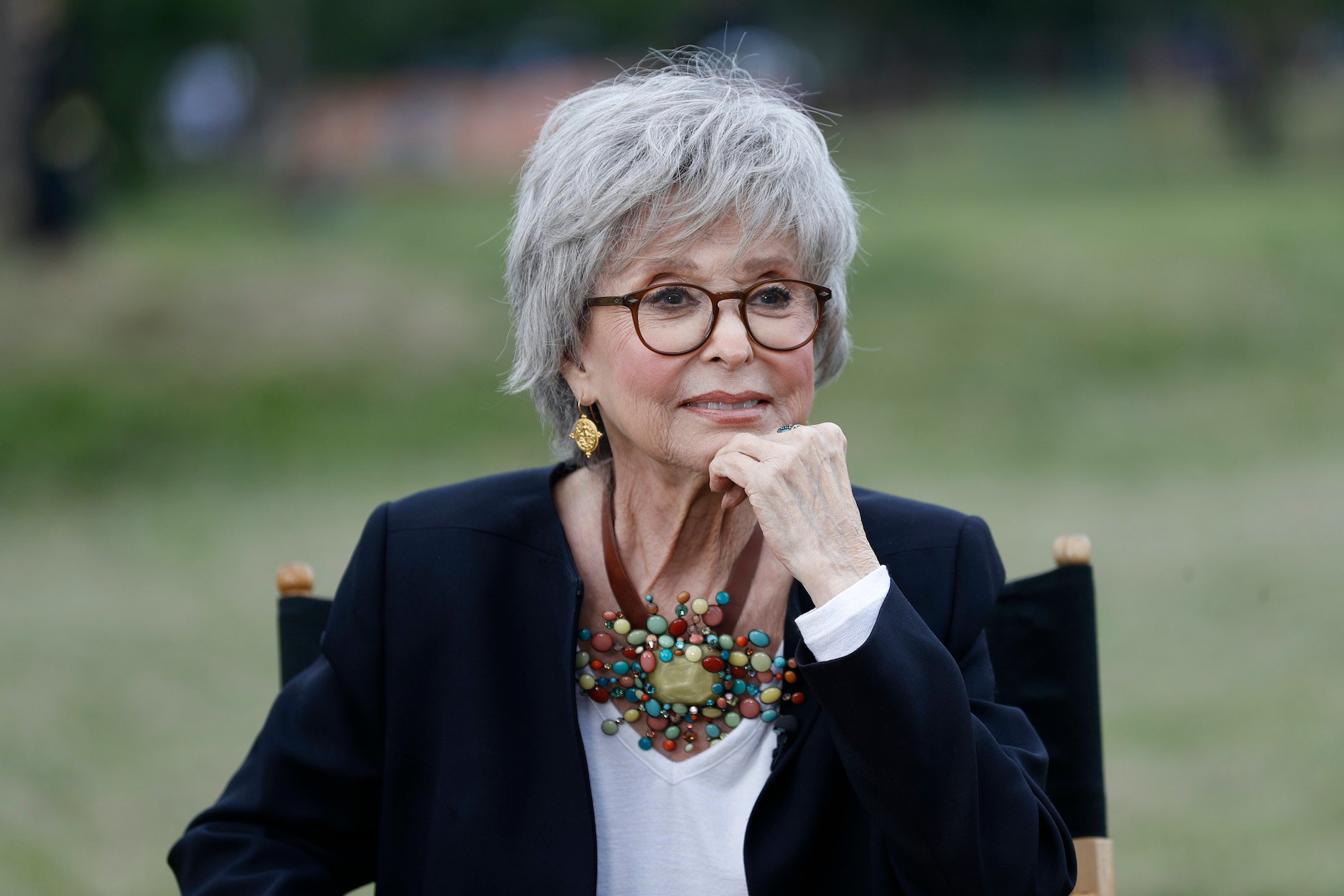 Rita Moreno at the Rita Moreno Puerto Rican Day Parade Celebration during the 2021 Tribeca Festival on June 13, 2021 in New York City. She sits in a chair on a lawn with her hand under her chin as she smiles.