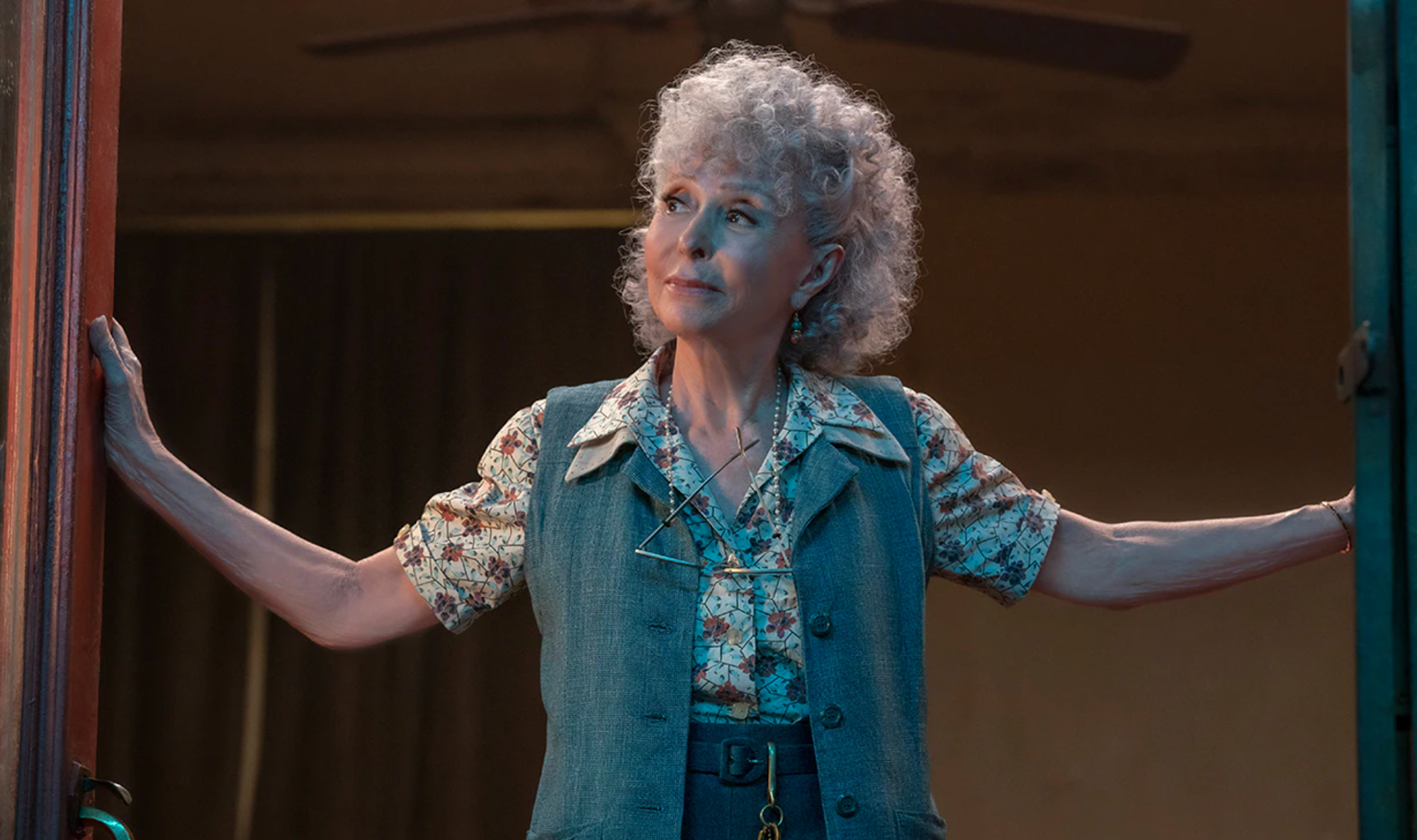 Rita Moreno as Valentina in Steven Spielberg's 'West Side Story.' She stands in a doorway with her arms on either side of the frame while wearing pants, a floral button-up shirt, and a grey vest.
