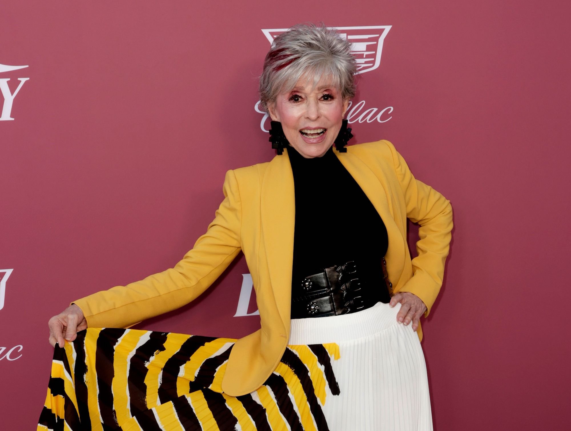 Honoree Rita Moreno attends Variety's Power Of Women event on Sept. 30, 2021 in California. She stands in front of a mauve backdrop wearing a black top, bright yellow suit jacket, and white, black, and yellow patterned skirt. She holds her skirt out to the side as she smiles for the camera on the event's red carpet.