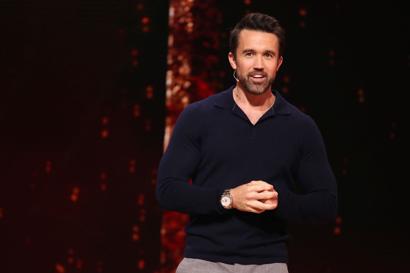 Rob McElhenney dressed in a long sleeve blue polo in front of a orange and black background.