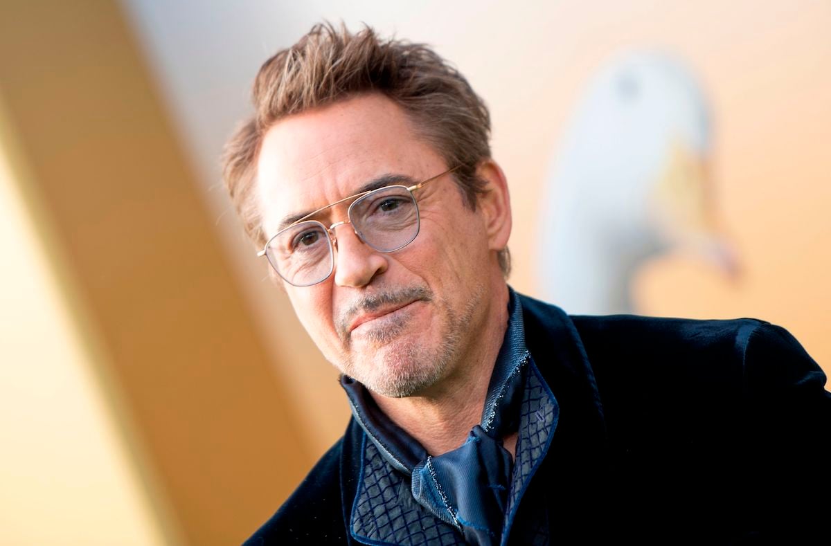 Robert Downey Jr smiling. He stands in front of a gold backdrop while wearing a navy blue velvet suit jacket and blue neck scarf and slightly blue-tinted glasses.