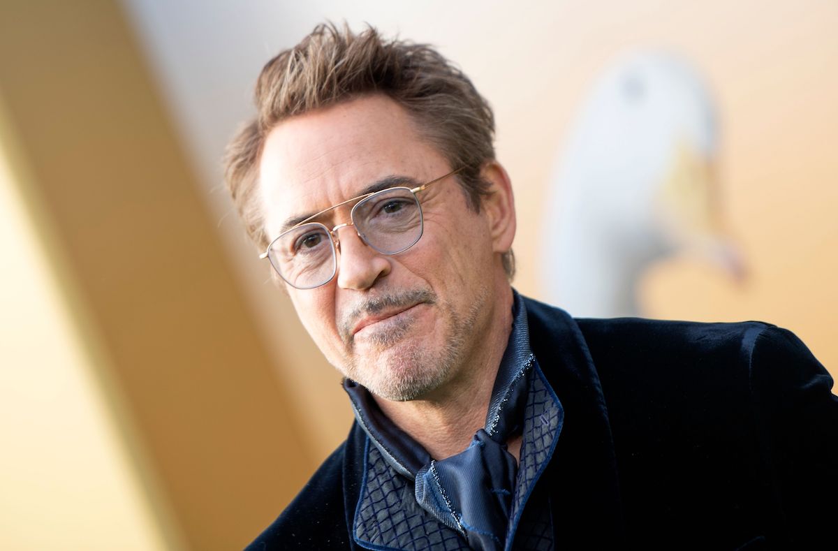 Robert Downey Jr smiling. He stands in front of a gold backdrop while wearing a navy blue velvet suit jacket and blue neck scarf and slightly blue-tinted glasses.