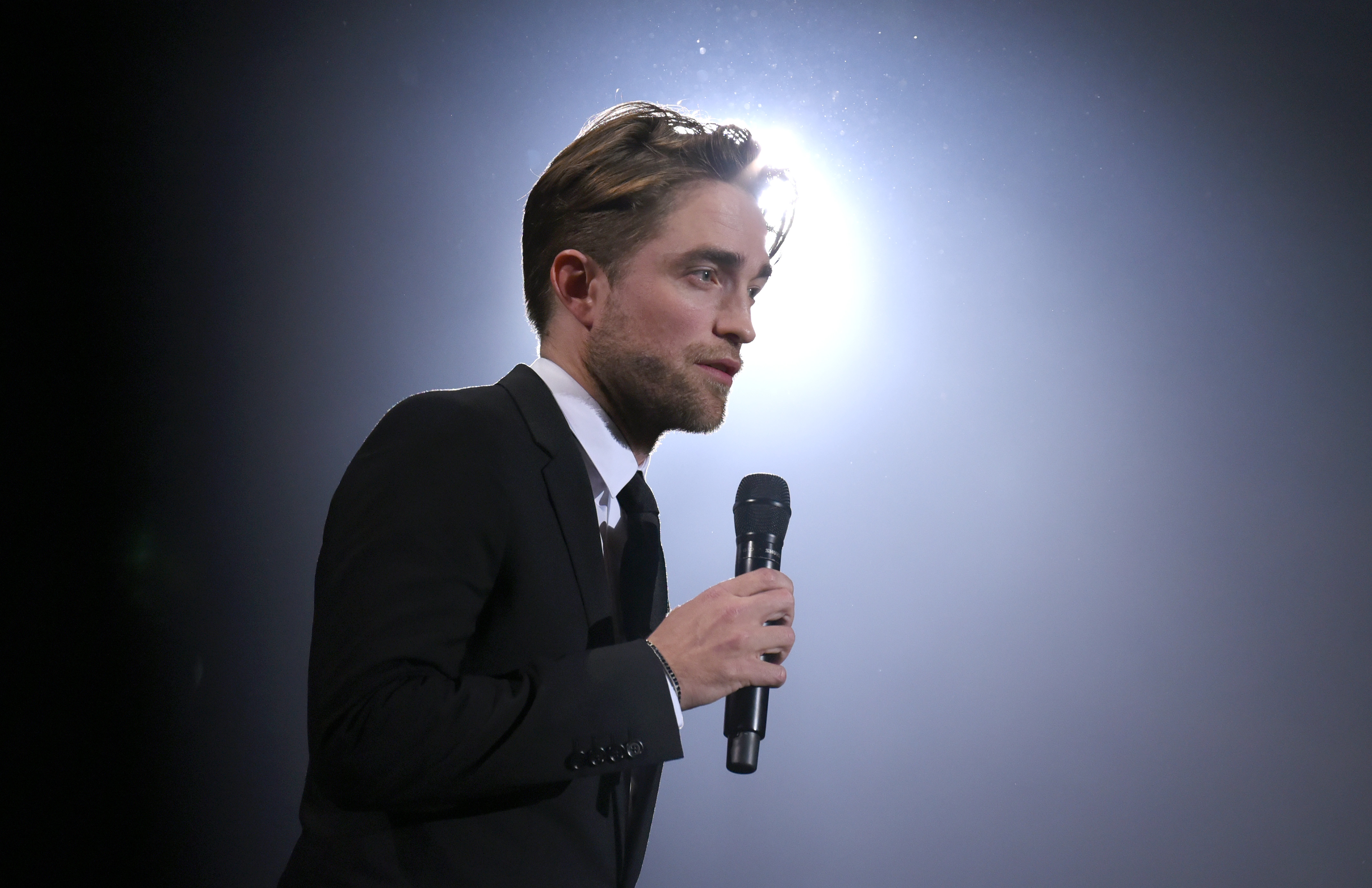 Robert Pattinson, who is featured in the DC FanDome 2021 trailer, wears a black suit and tie and holds a microphone.