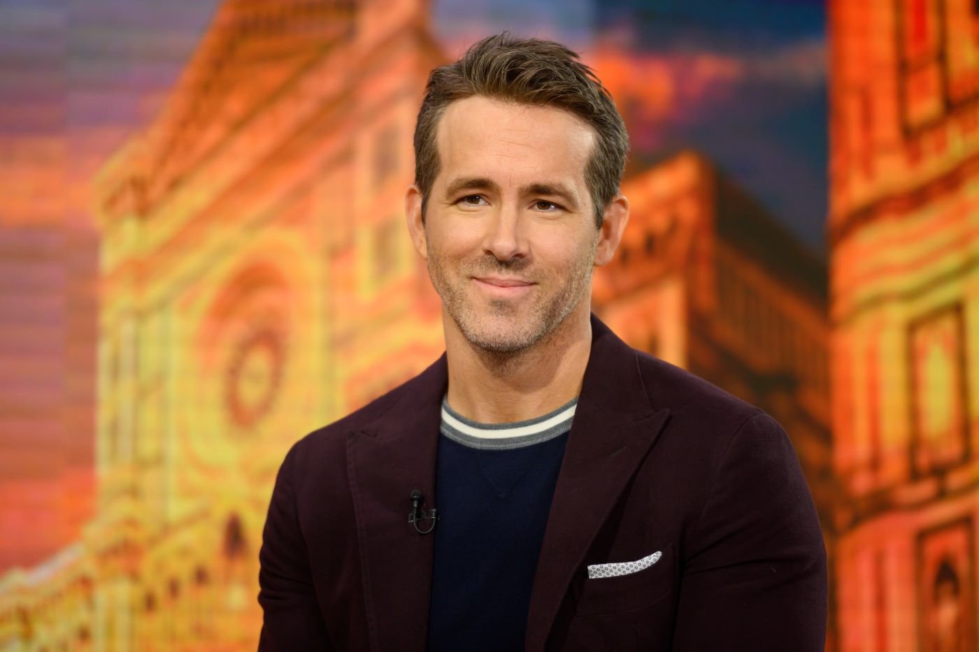 Ryan Reynolds dressed in a maroon suit with a black shirt underneath in front of a background of a digital building with an orange tint.